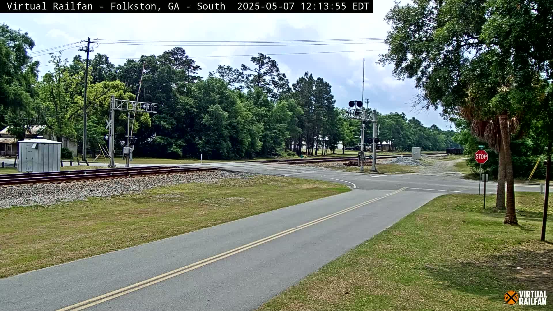 A paved road intersects with a railroad crossing under a partly cloudy sky, surrounded by green trees and grass.