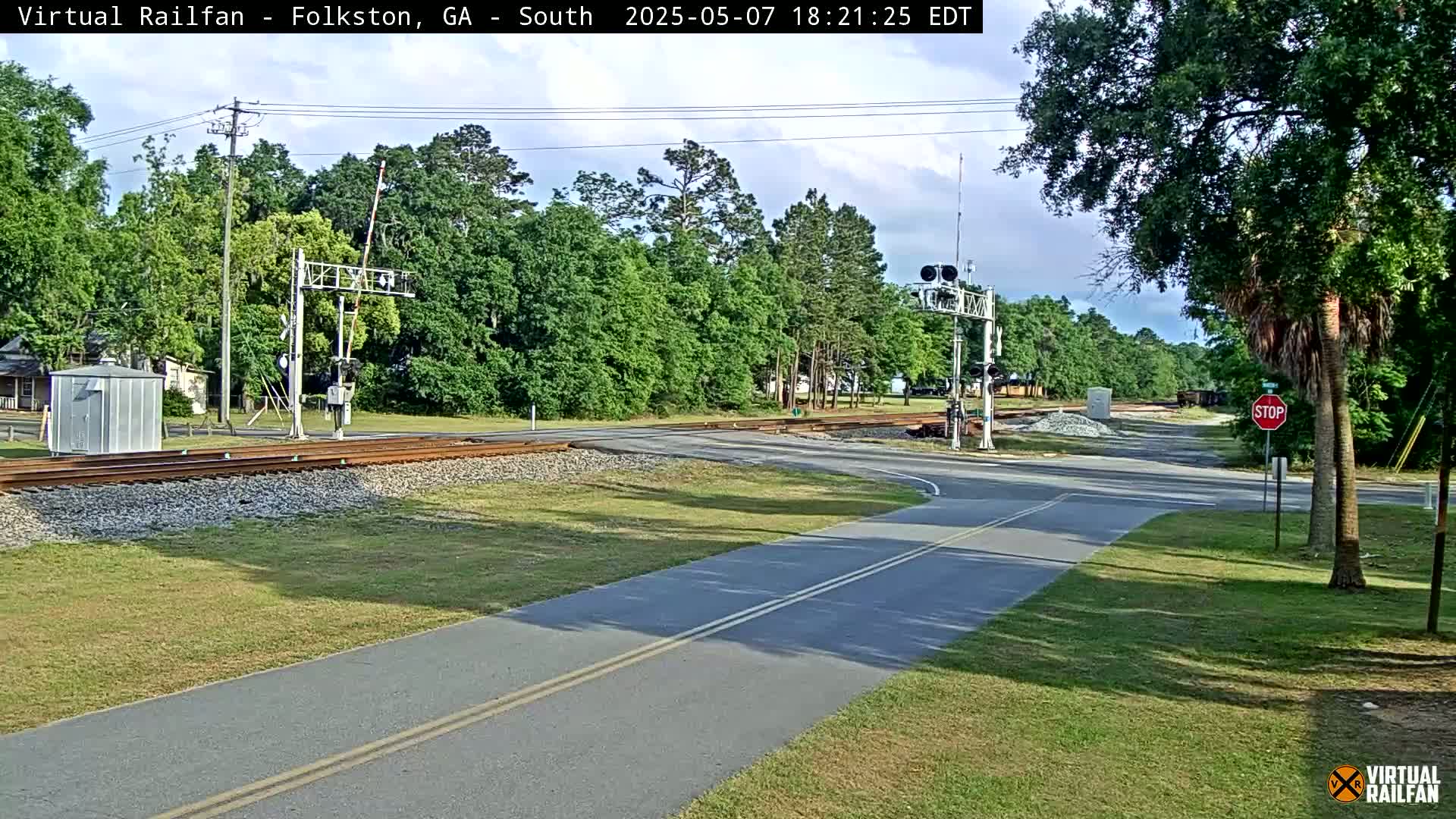 A mostly sunny view of a railroad crossing with a road and a paved path intersecting, surrounded by green trees and grass.