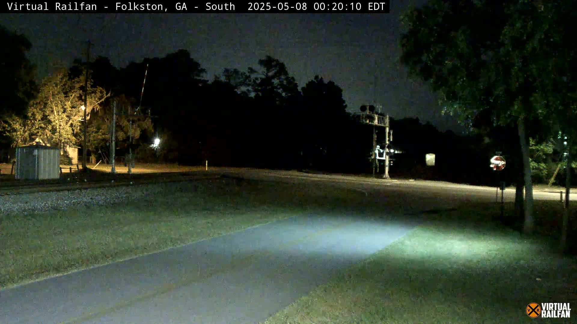 A nighttime view of a paved path intersecting with a road near a railroad crossing, surrounded by trees and under a dark sky.