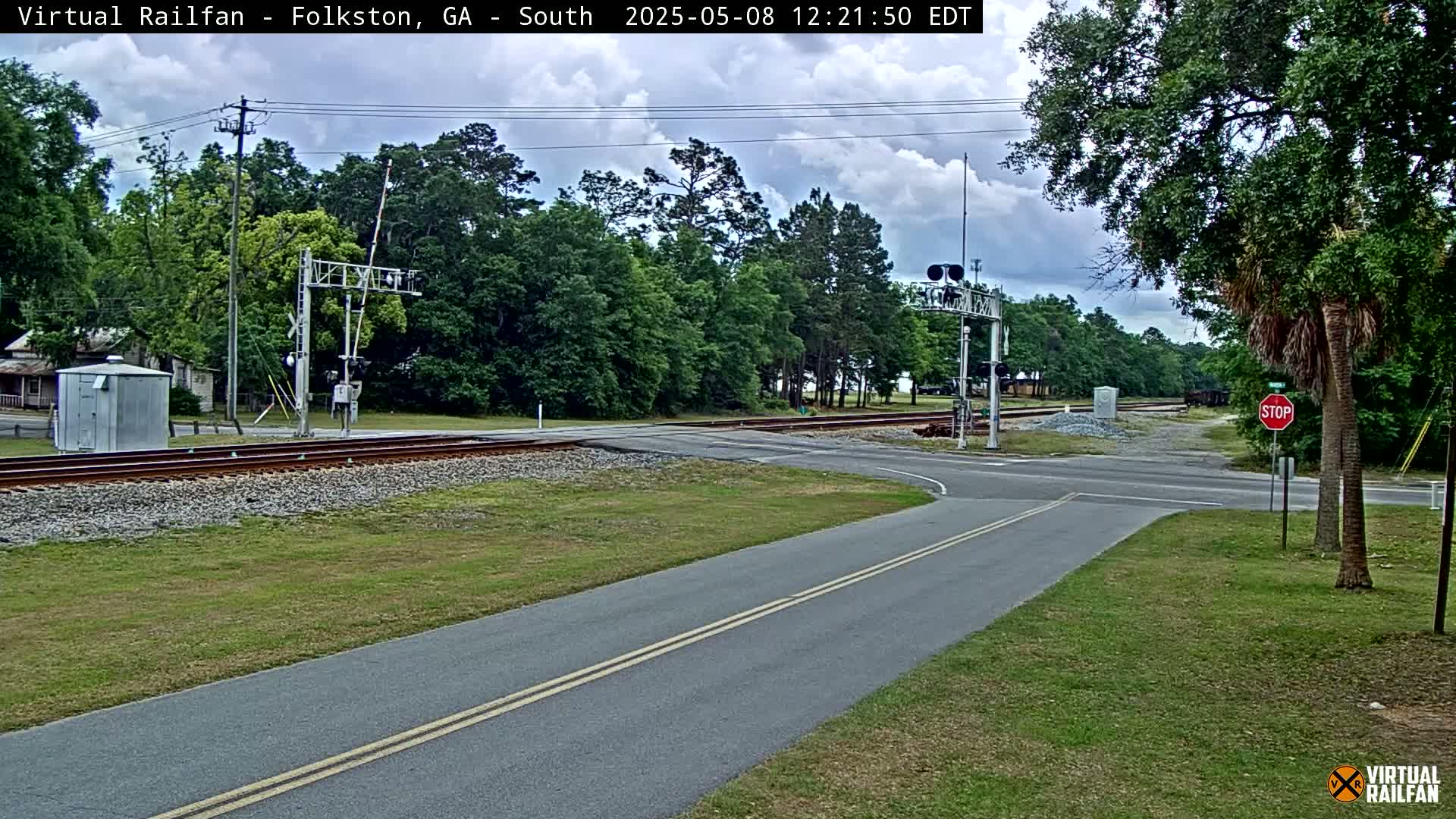 A partly cloudy day shows a railroad crossing with a road intersecting the tracks, surrounded by green grass and trees.