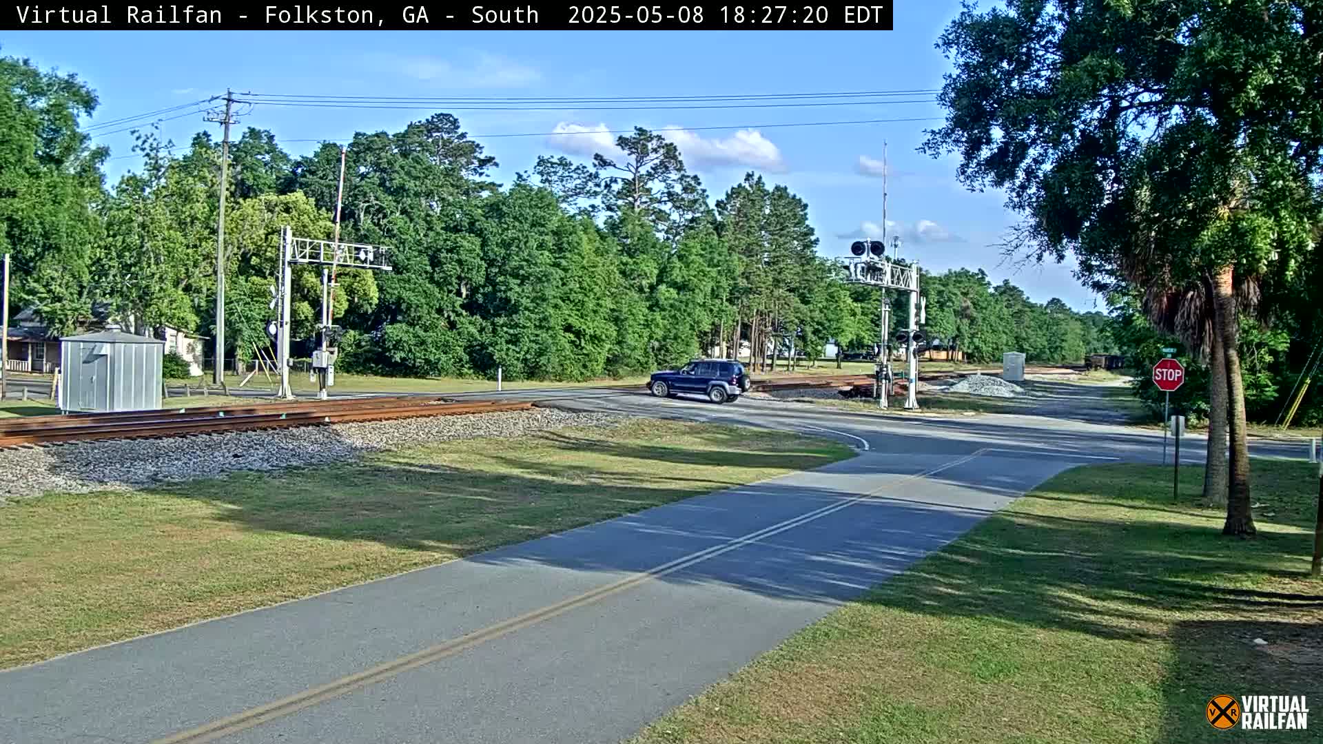 A dark-colored SUV drives past a railroad crossing on a sunny day, with trees lining the road and a paved path visible in the foreground.