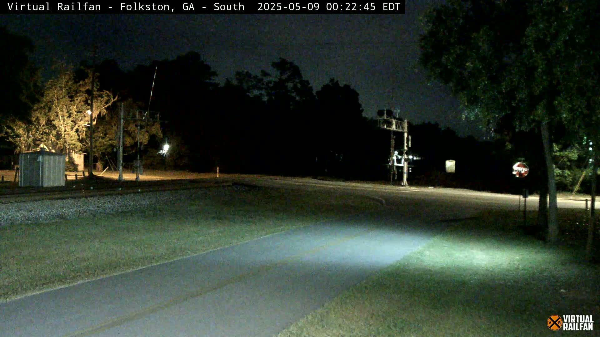 A nighttime view of a railroad crossing with a paved path and surrounding trees under a dark sky.