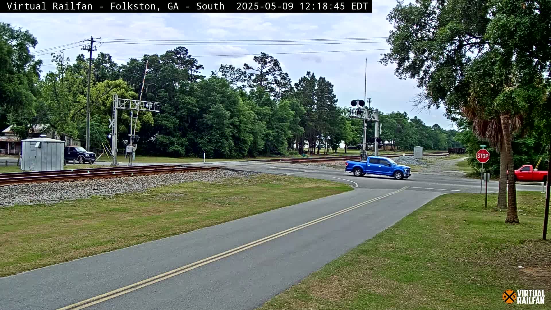 A blue pickup truck is turning right onto a road that crosses railroad tracks, under a partly cloudy sky, with lush green trees and other vehicles visible.