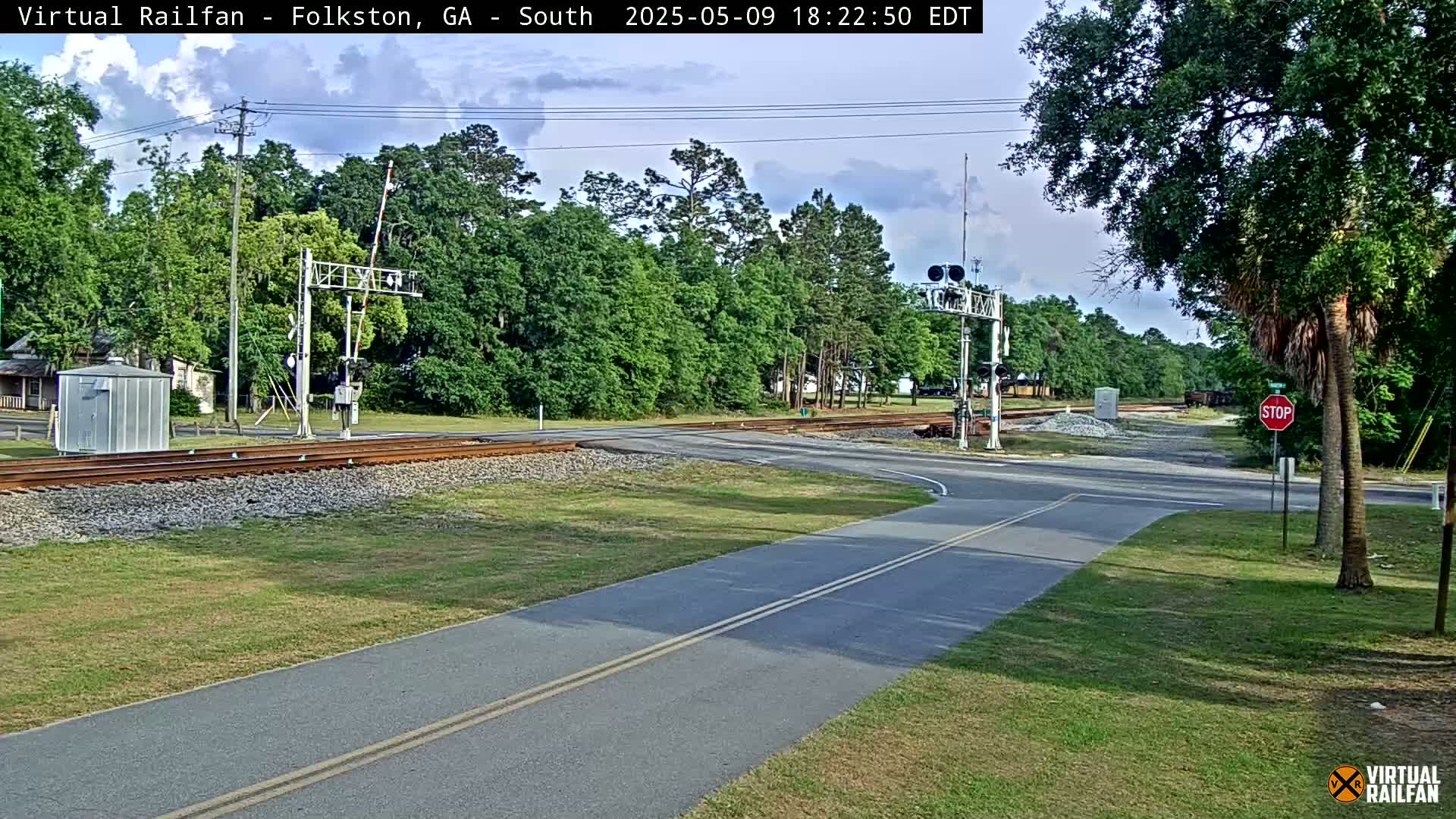 A paved road intersects with a railroad crossing under a partly cloudy sky, surrounded by green trees and grass.