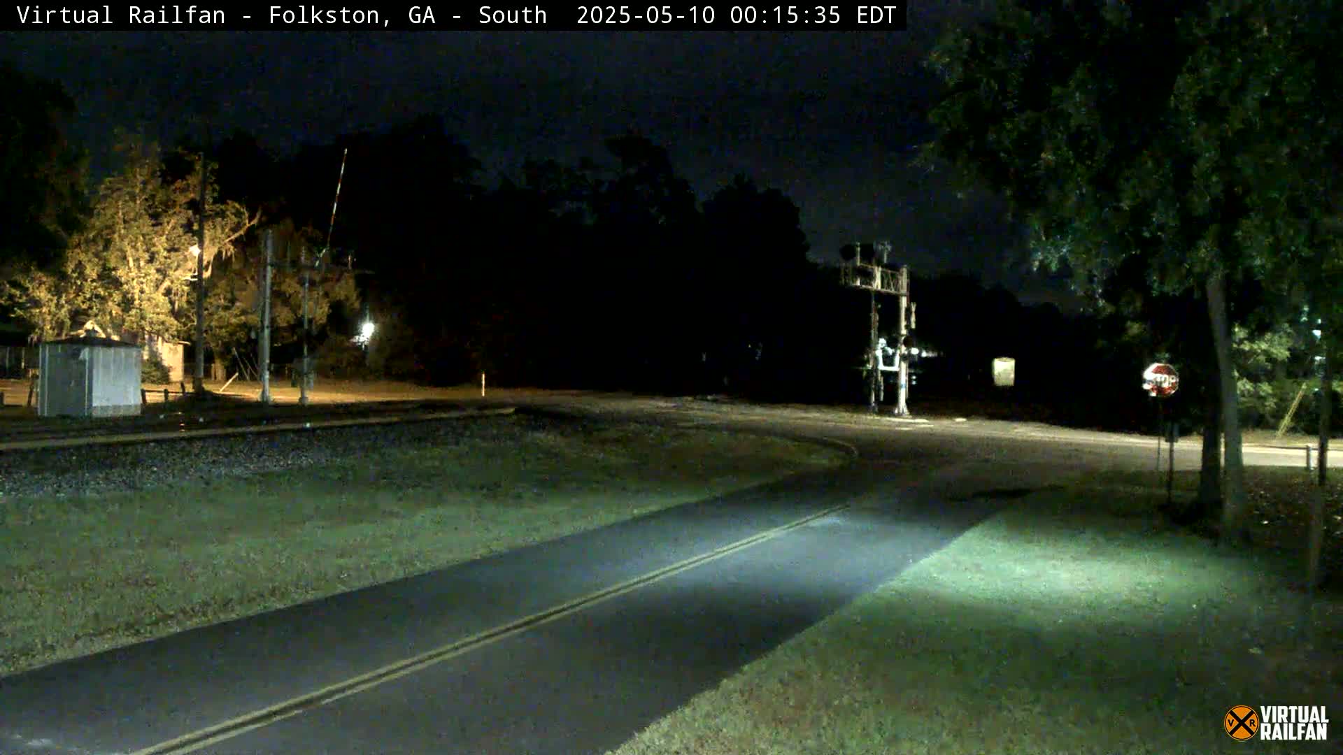 A nighttime view shows a paved road intersecting with a railroad crossing, surrounded by trees and grass.