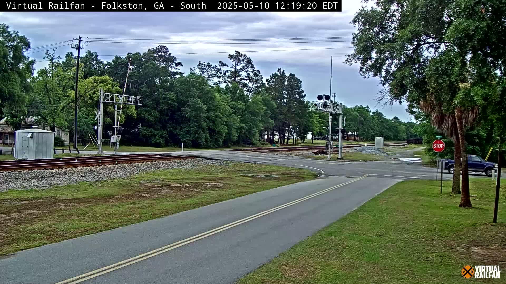 A road intersects with railroad tracks under a partly cloudy sky, with trees lining the area and a vehicle stopped at a stop sign.