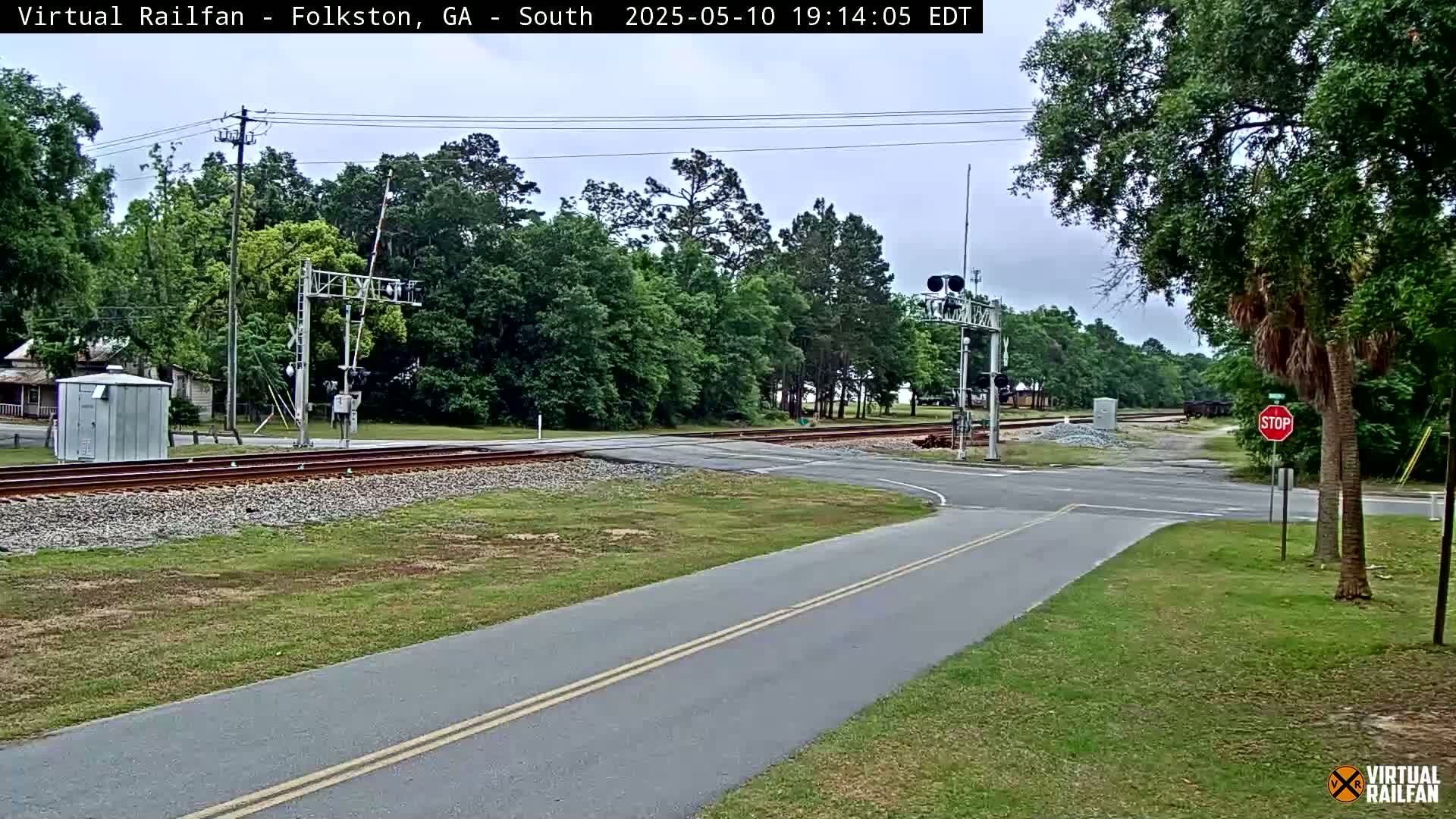 A paved road intersects with a railroad crossing under a mostly cloudy sky, surrounded by green trees and grass.