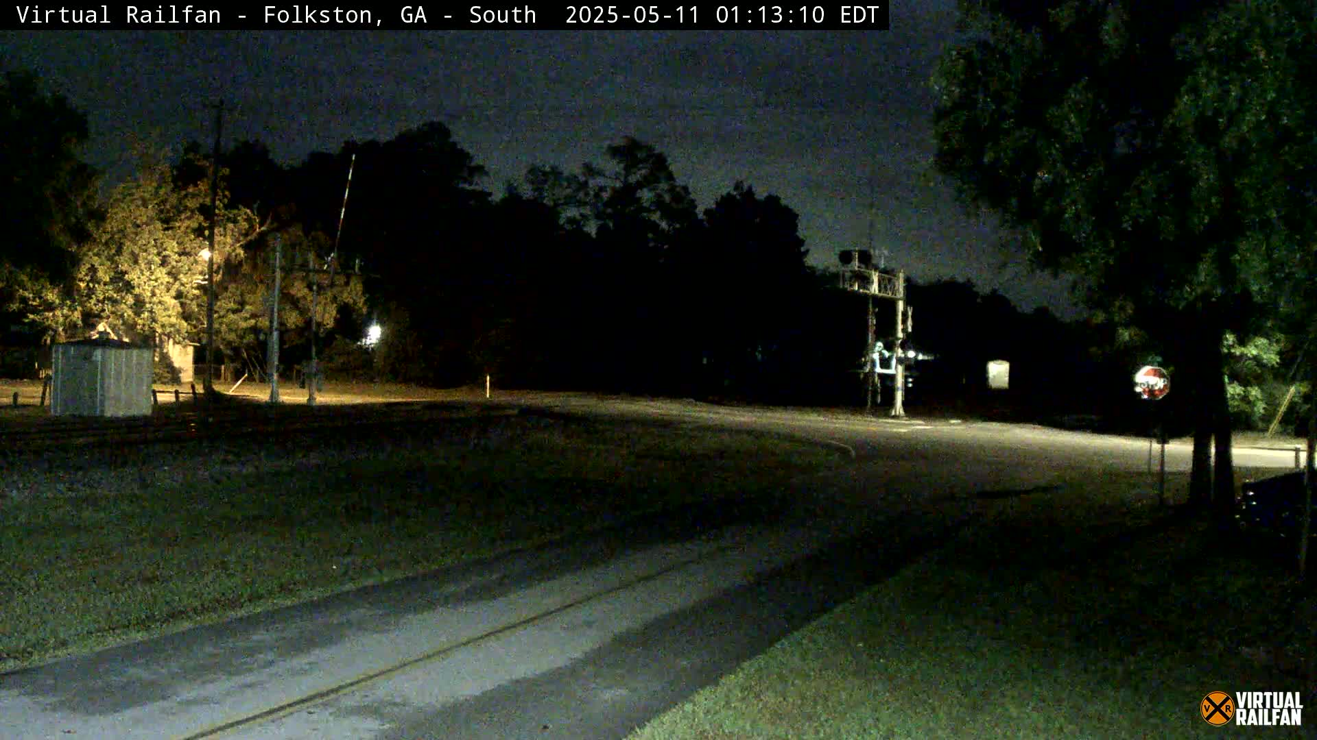 A nighttime view of a road intersecting with railroad tracks, featuring a railway crossing signal and surrounding trees under a dark sky.