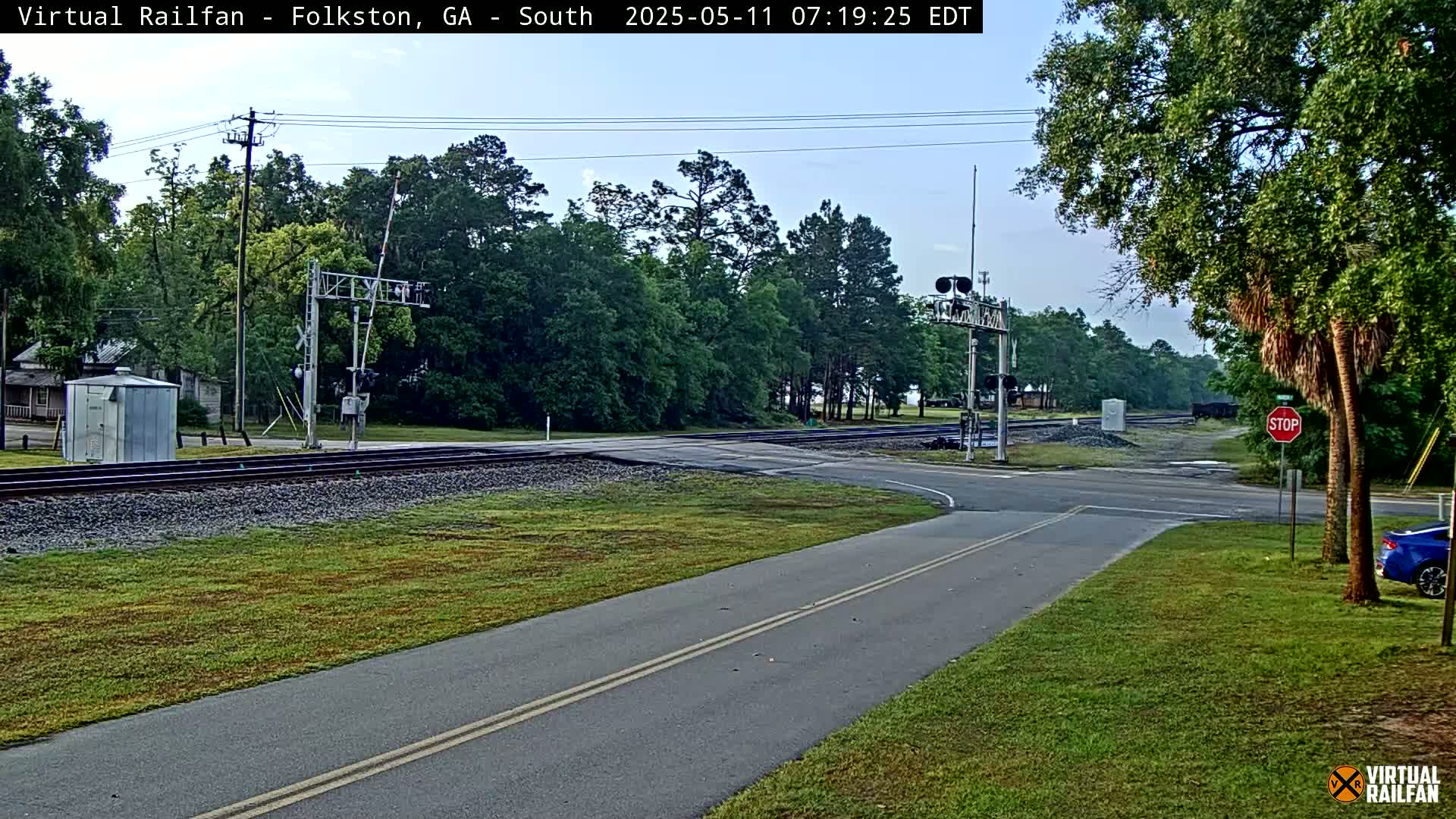 A road intersects a railroad crossing under a partly cloudy sky, with trees and a small building visible in the background.