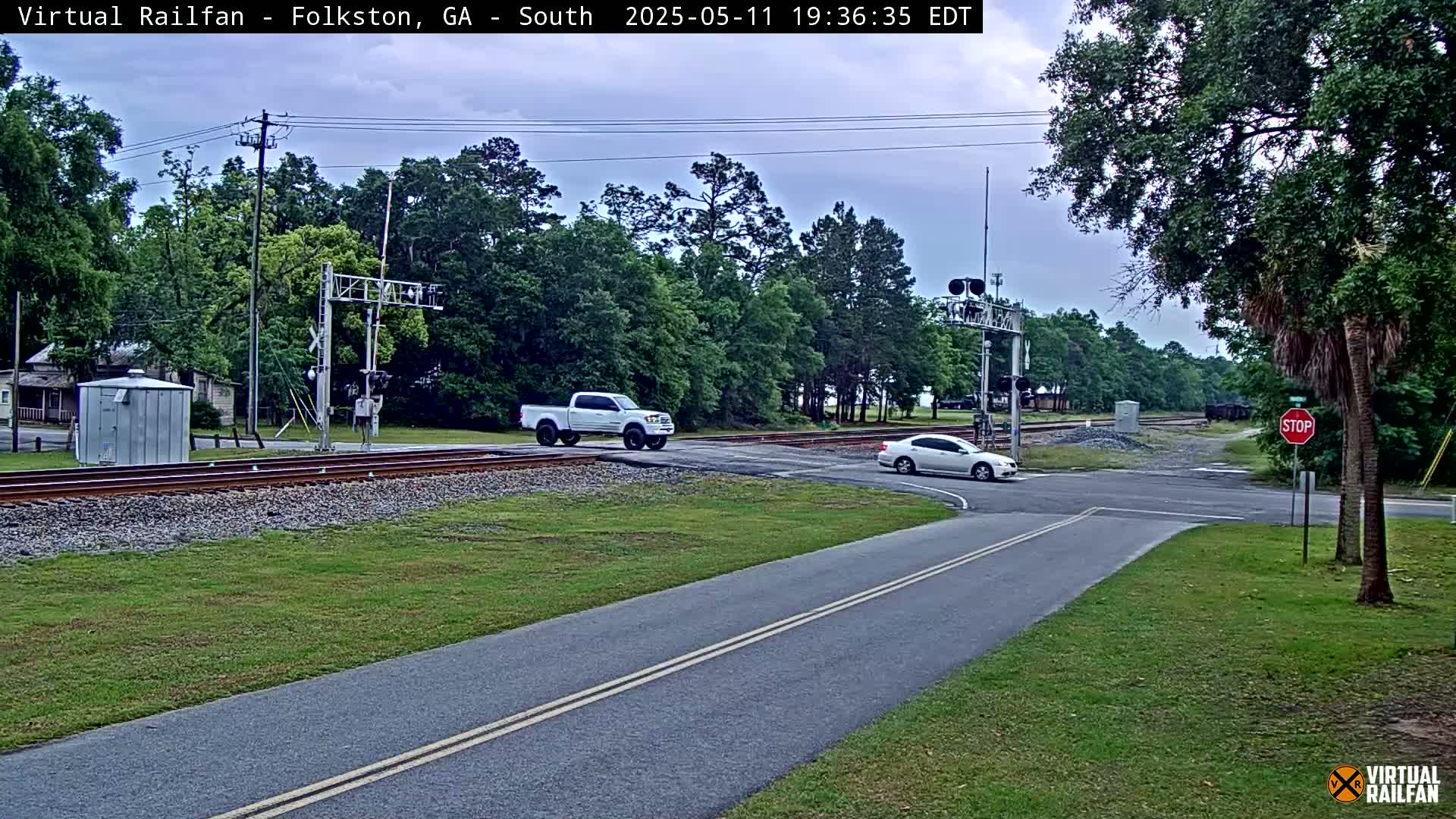 A mostly cloudy day shows a road intersecting with railroad tracks, where a white pickup truck and a white car are crossing.