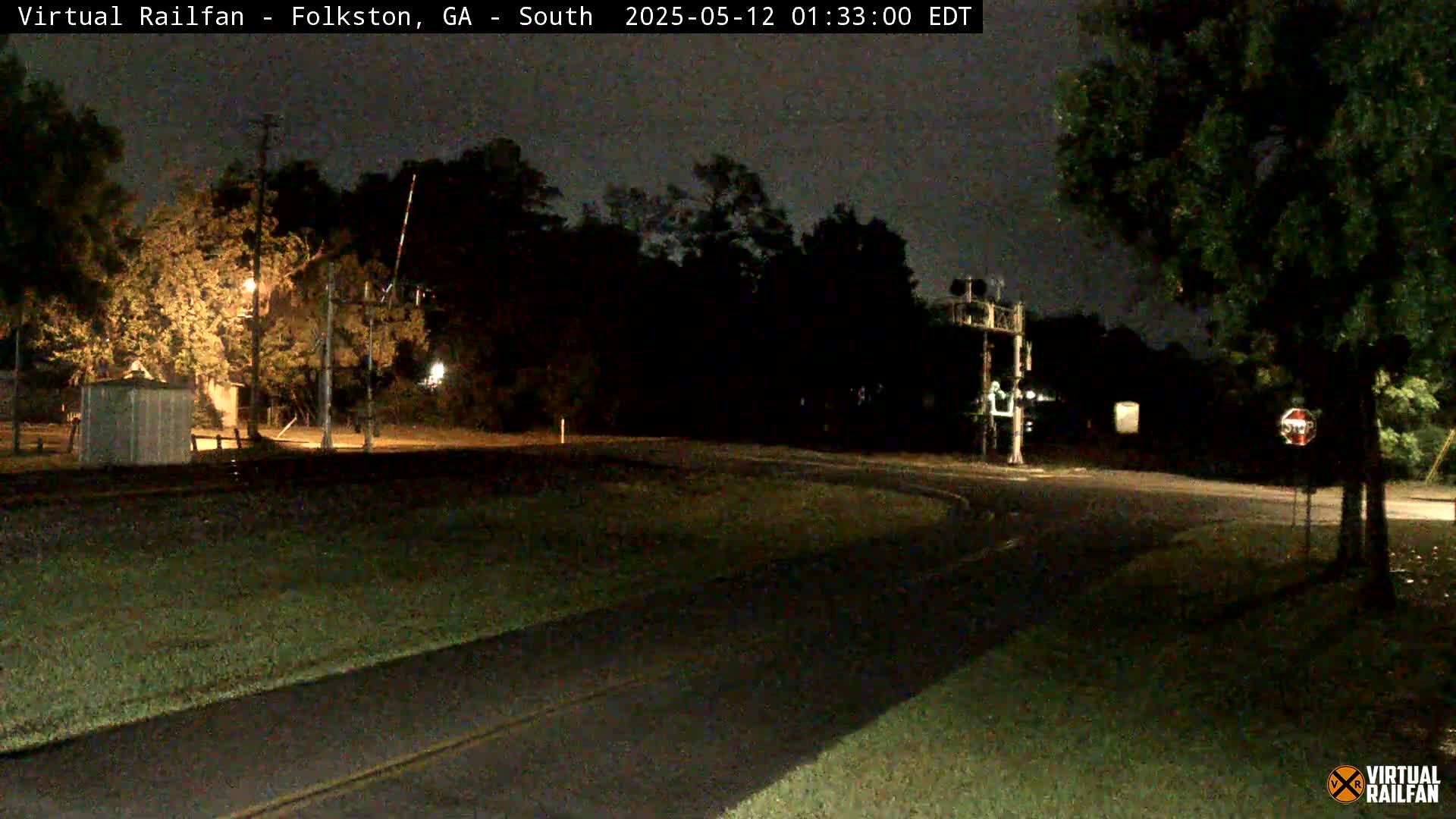 A nighttime view of a dark, empty road curving past a railroad crossing with trees and a small building visible under dim lighting.