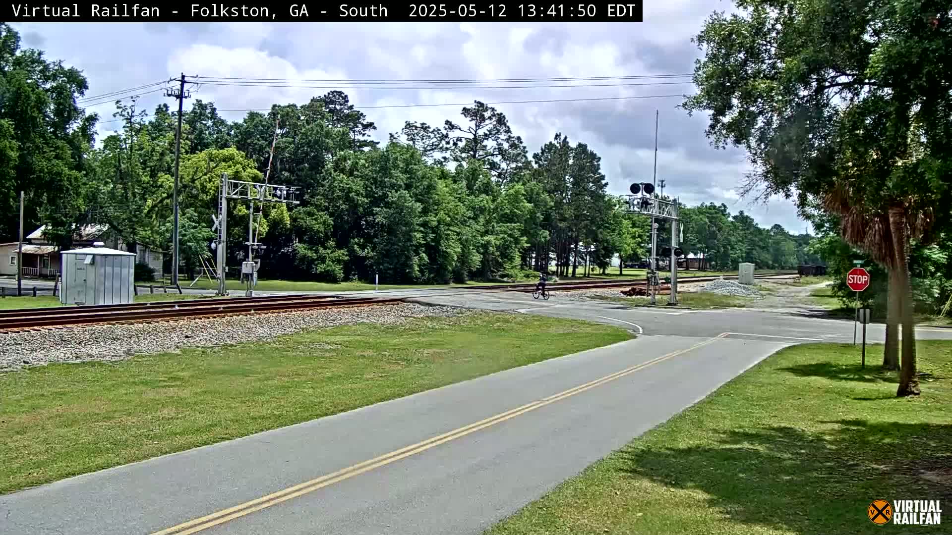 A cyclist rides across a road near railroad tracks on a partly cloudy day.