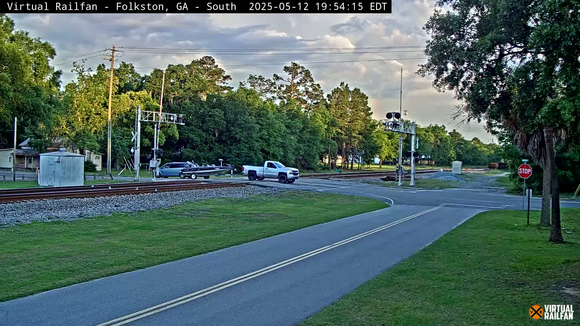 A partly cloudy evening scene shows a road intersecting railroad tracks, with a pickup truck and a vehicle towing a boat stopped near the crossing.