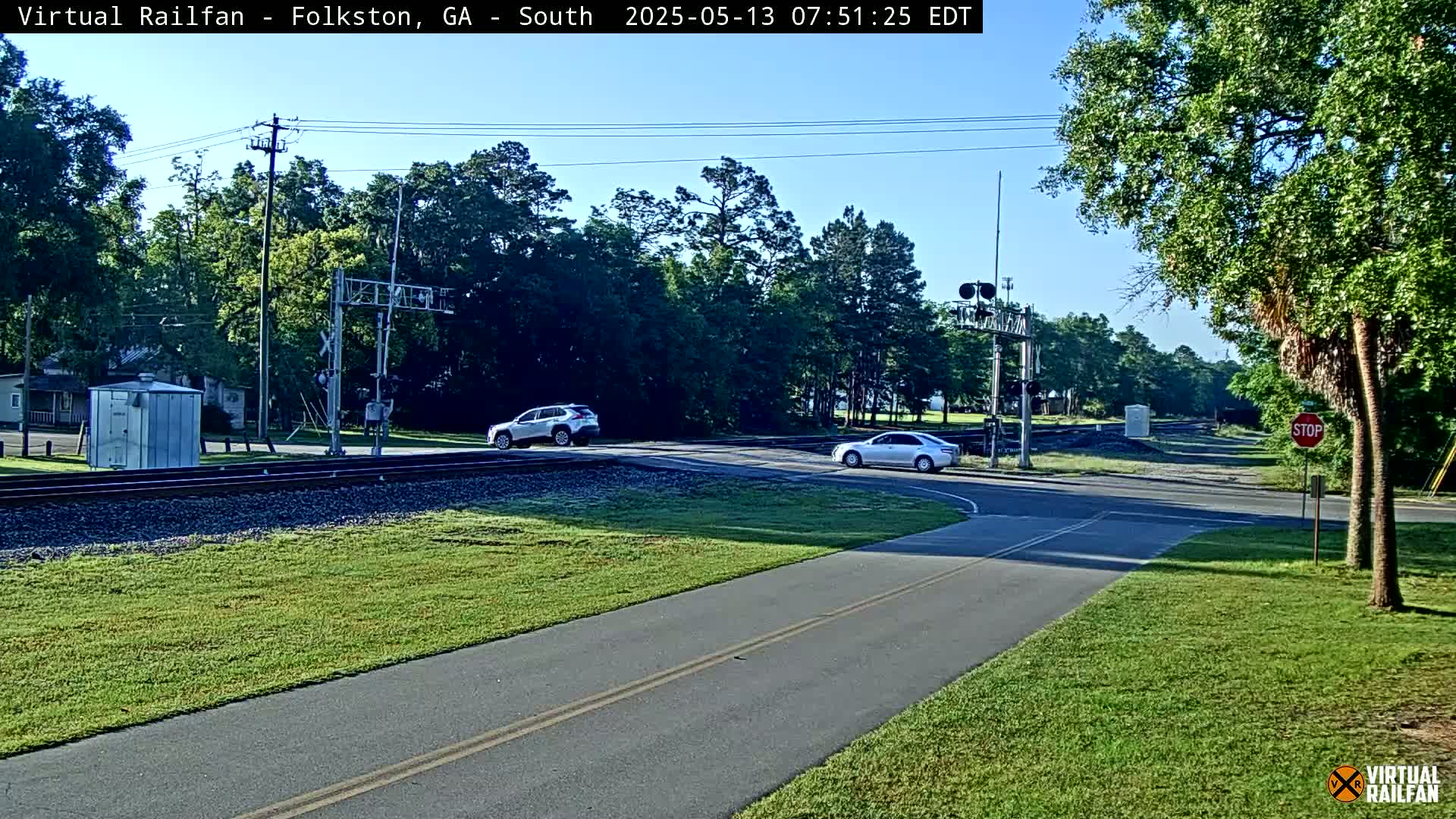 A sunny day shows a railroad crossing with two cars stopped near a paved path; trees and grass surround the area.
