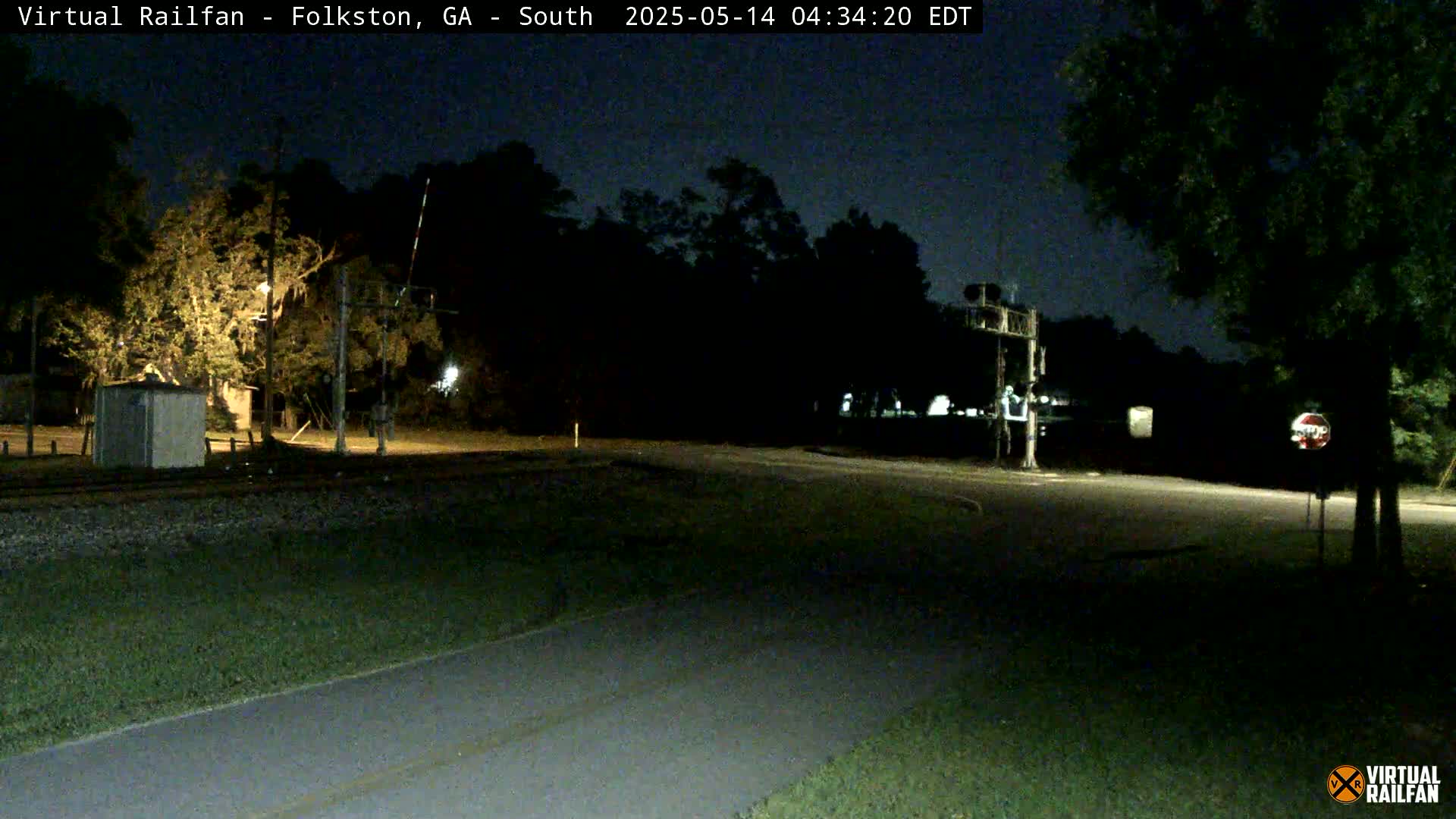A nighttime view of a rural road intersection with railroad tracks, trees, and a small building under a dark sky.