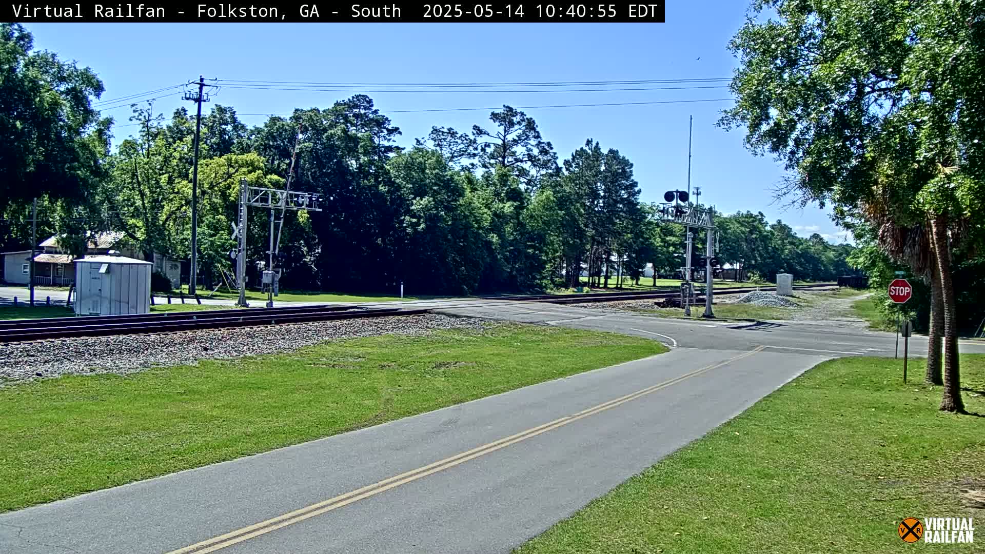 A sunny day reveals a railroad crossing with a road intersecting the tracks, surrounded by green grass and lush trees.