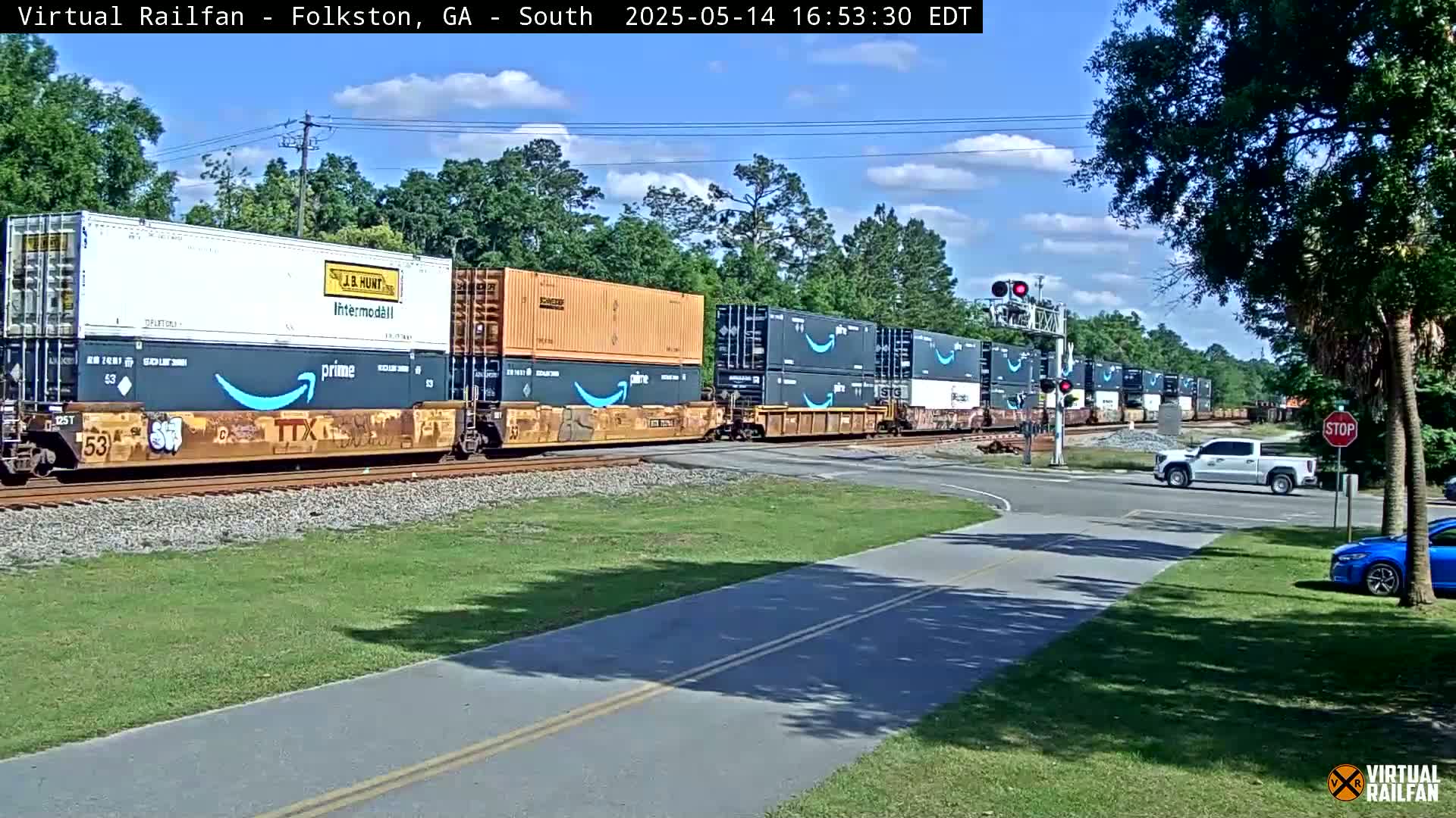 A long freight train carrying shipping containers, some displaying the Amazon logo, passes a road intersection under a partly cloudy sky on a sunny day.