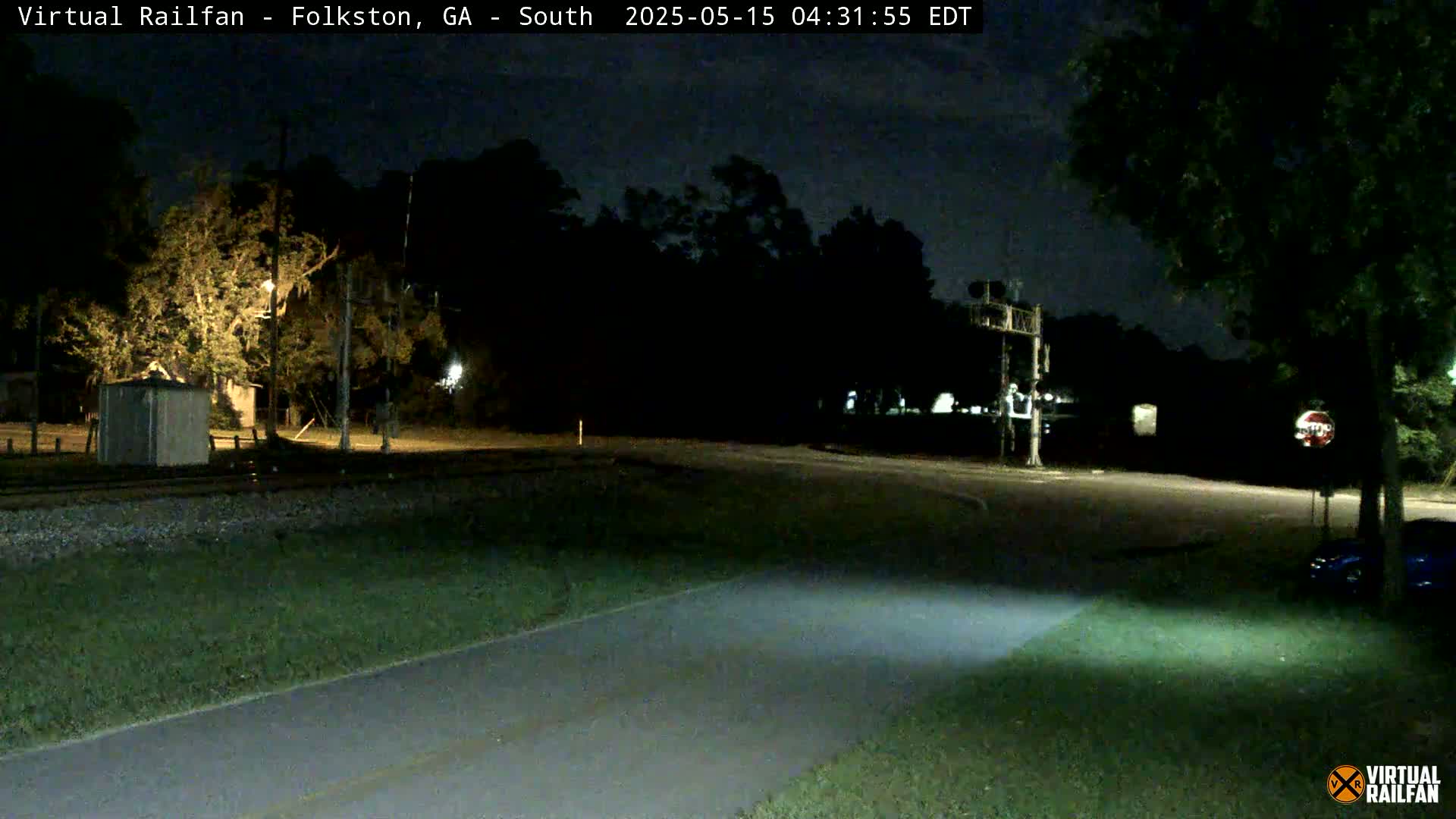 A nighttime view of a road intersecting railroad tracks, with a stop sign and some trees visible under a dark sky.