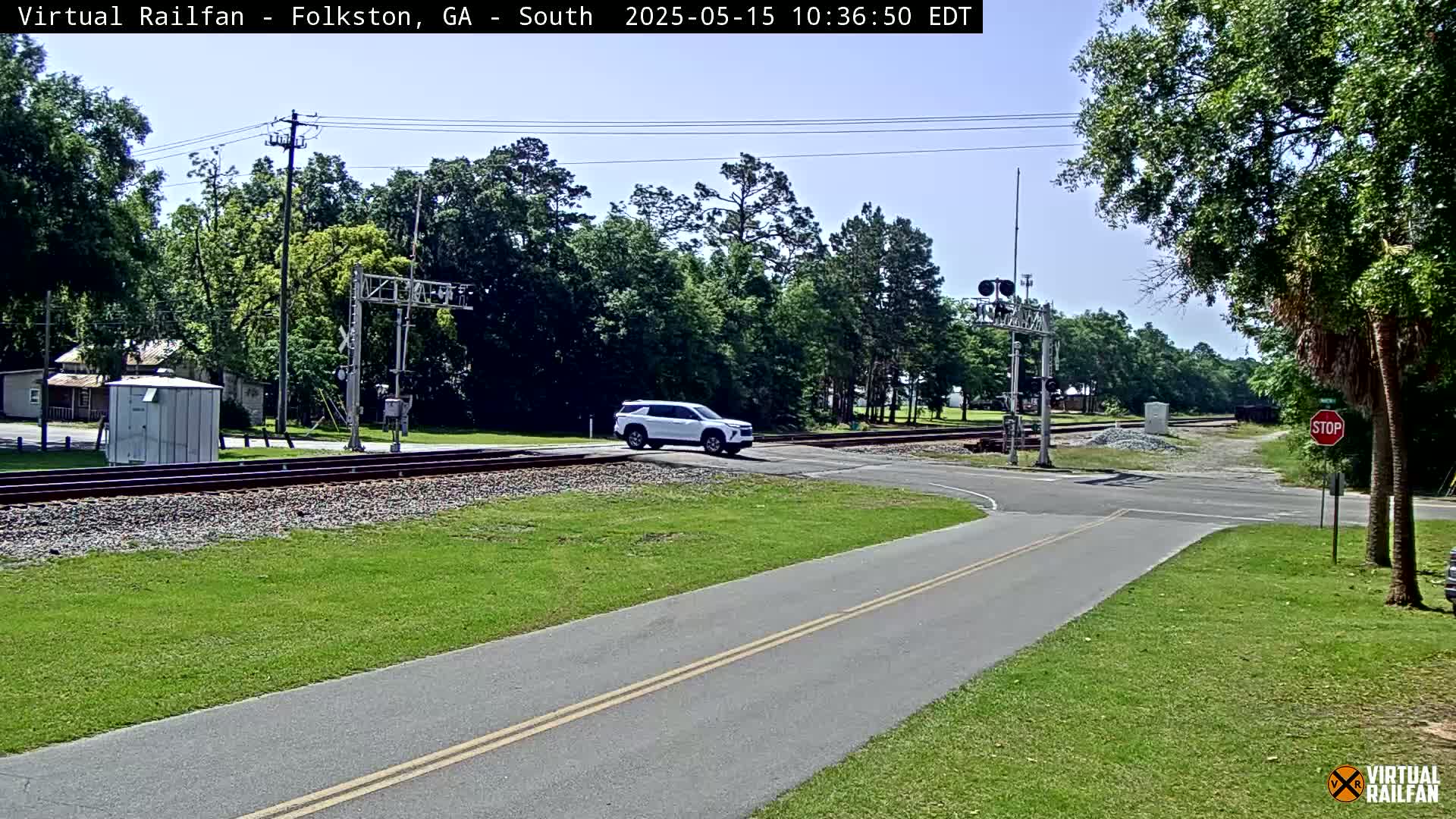 A white SUV crosses a railroad crossing on a sunny day, with trees and grass lining the road and railroad tracks.