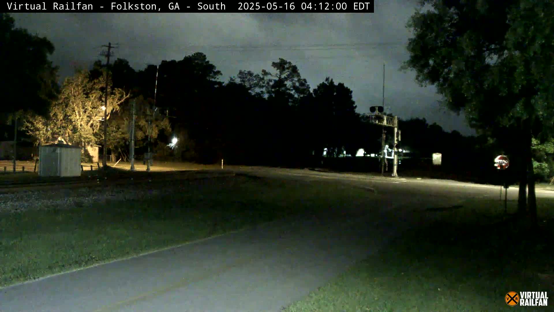 A nighttime view of a road intersection near a railroad crossing, surrounded by trees under a dark sky.