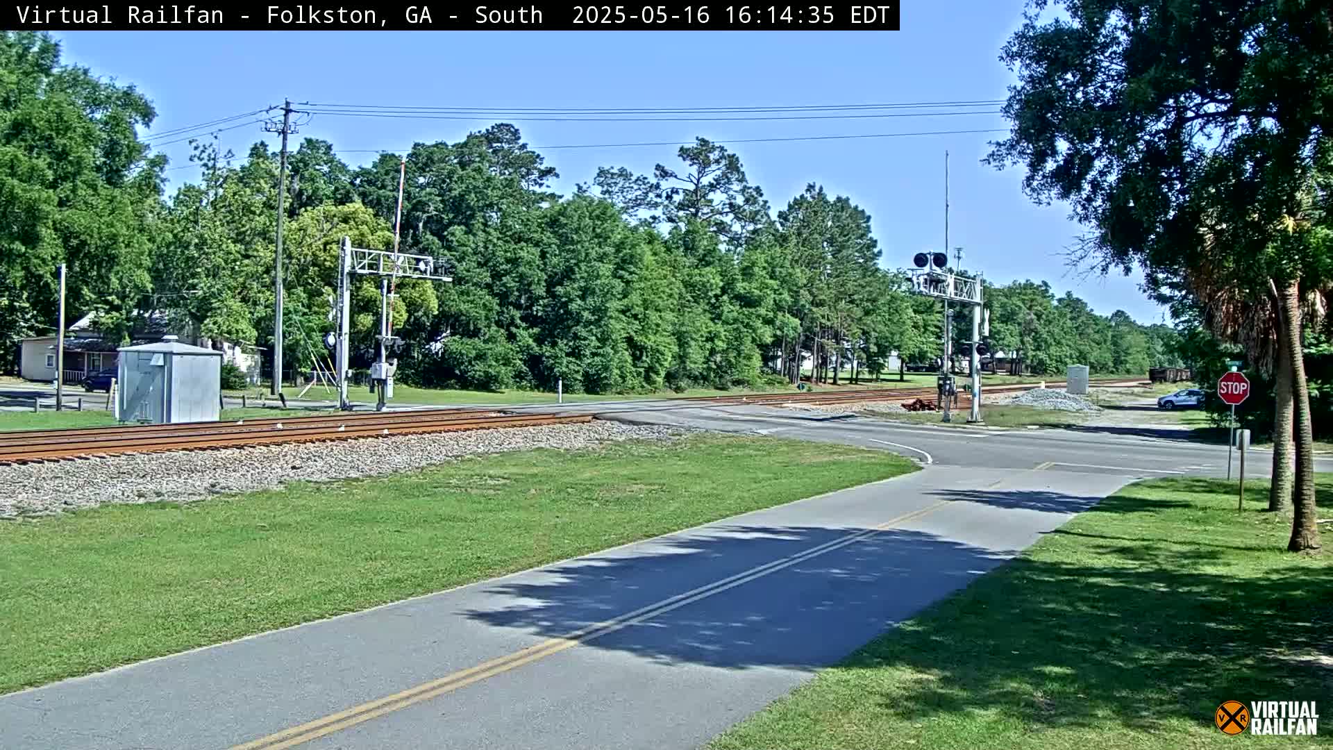 A sunny day shows a railroad crossing with a road intersecting it, surrounded by green grass and trees.