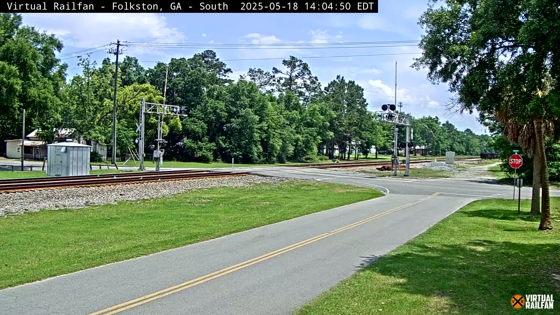 A sunny day reveals a railroad crossing intersecting a paved road, surrounded by lush green grass and trees.