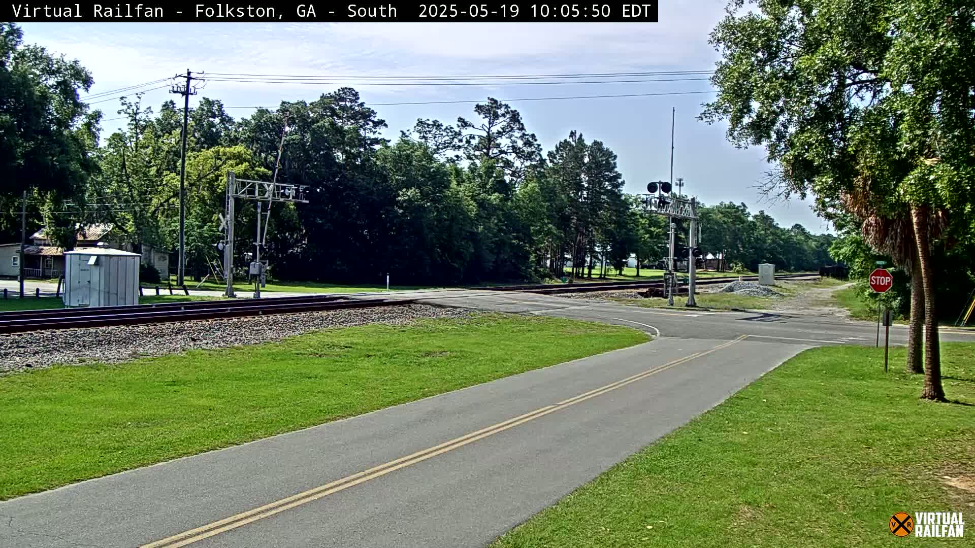 A paved road intersects a railroad crossing under a mostly sunny sky, surrounded by green grass and trees.