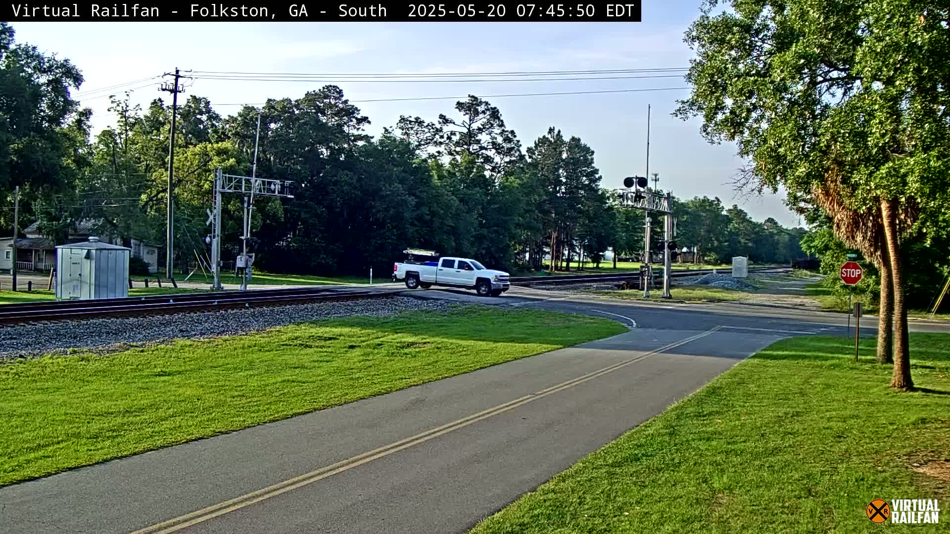 A white pickup truck is crossing a railroad crossing under a partly cloudy sky, with lush green grass and trees surrounding the area.