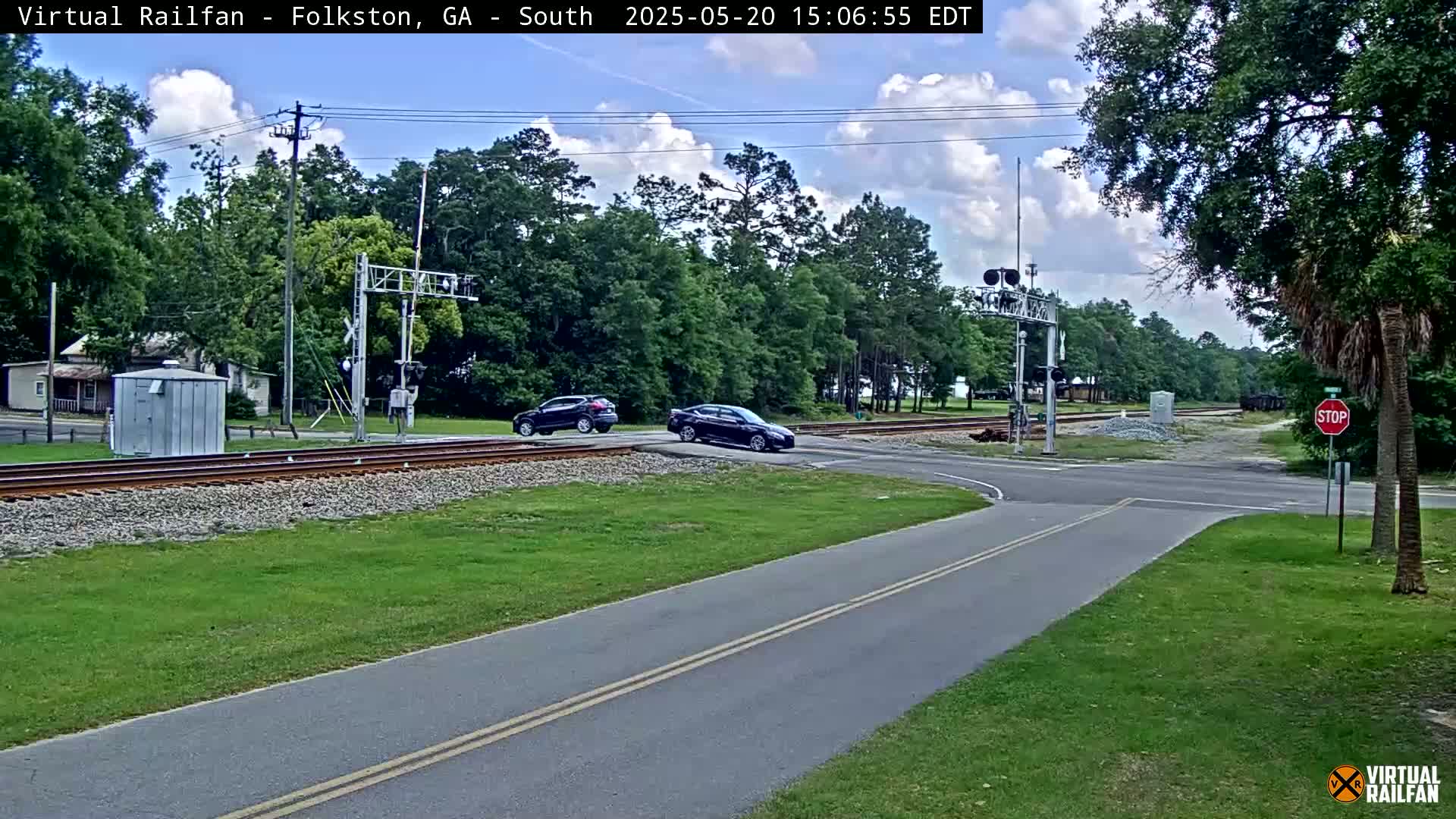 A partly cloudy day shows two dark-colored cars stopped at a railroad crossing, with a small building and lush green grass visible near the tracks.