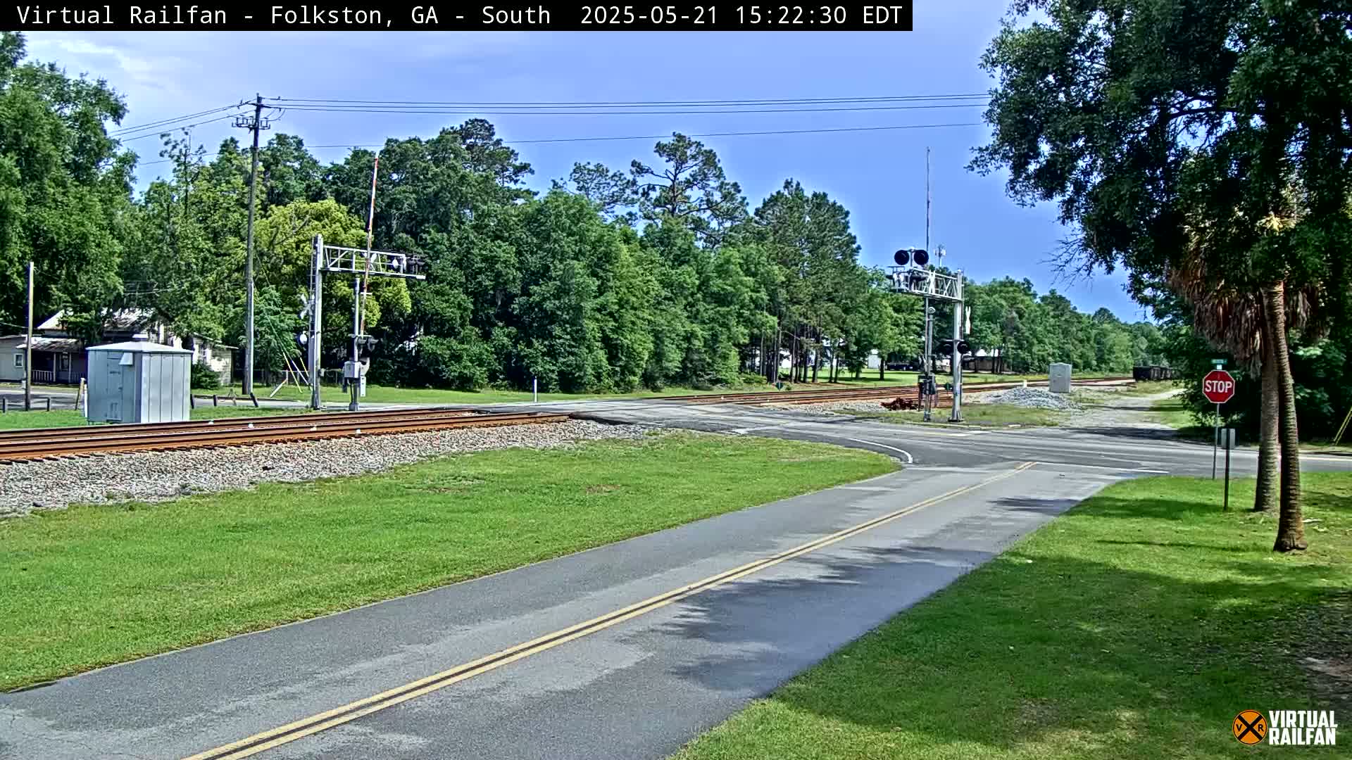 A sunny day reveals a railroad crossing with a road intersecting the tracks, surrounded by lush green grass and trees.