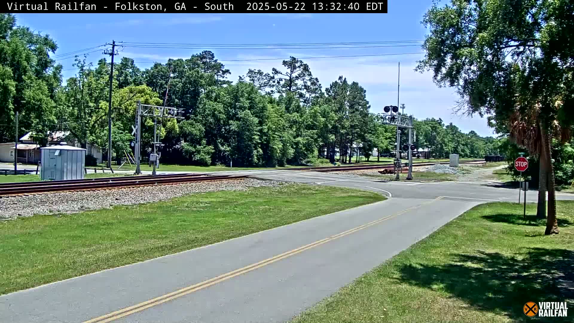 A paved road intersects with railroad tracks at a crossing under a mostly sunny sky, surrounded by lush green vegetation and trees.