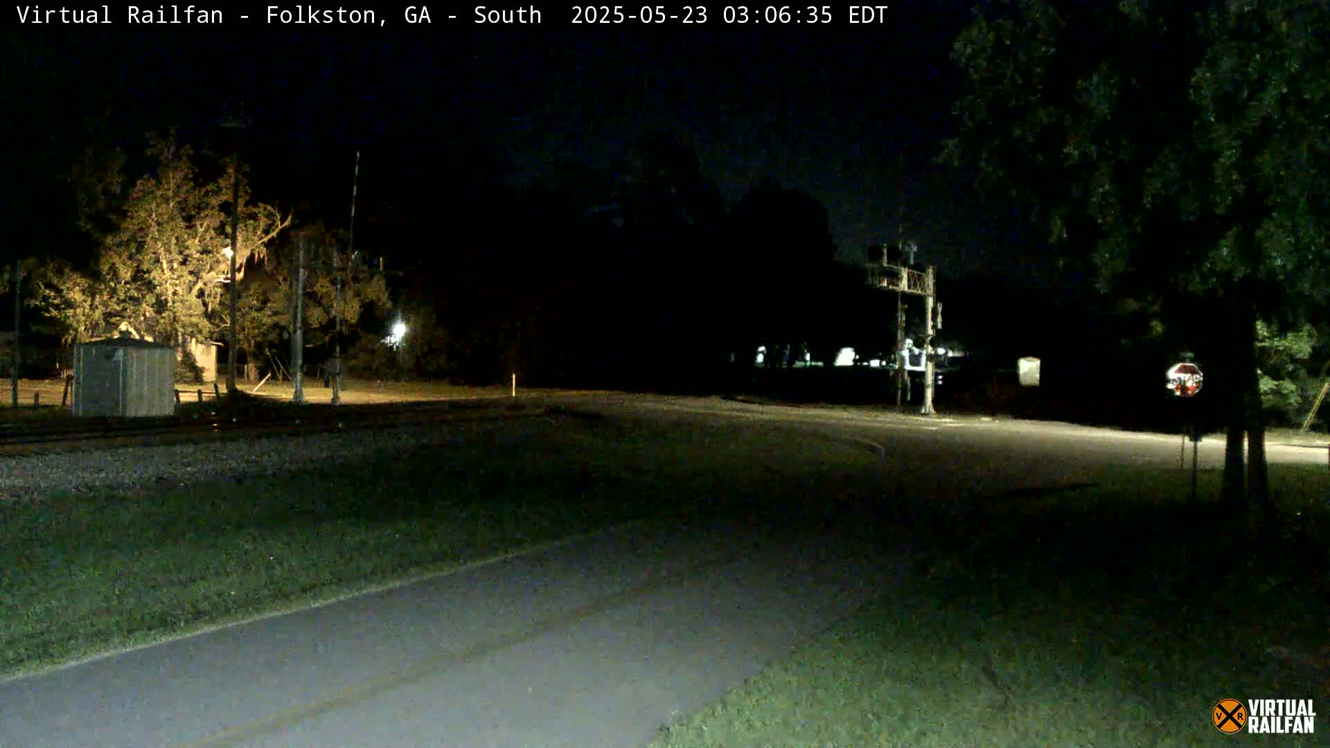 A nighttime view of a road intersection near a railroad crossing, with trees and a small building visible under dim lighting.