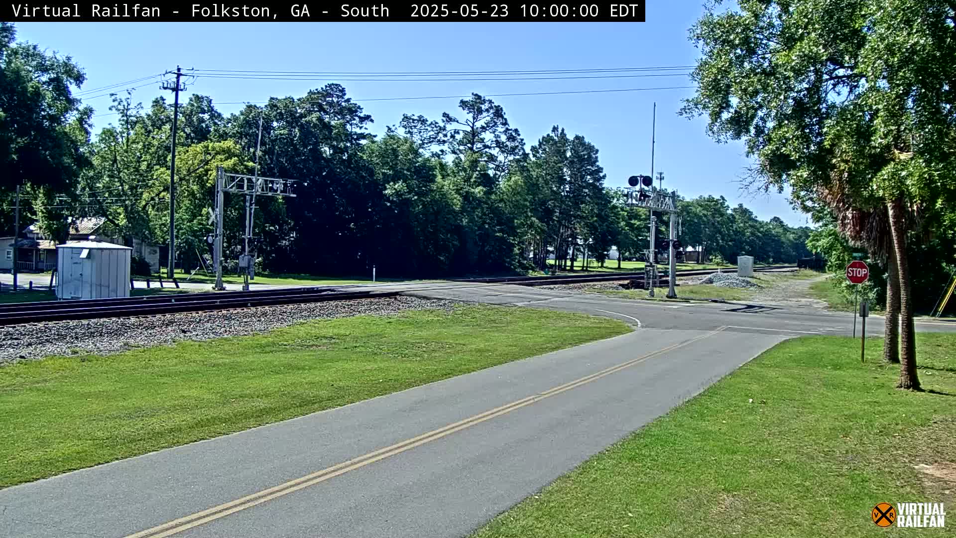 A paved road intersects with railroad tracks under a clear sky, surrounded by green grass and trees.