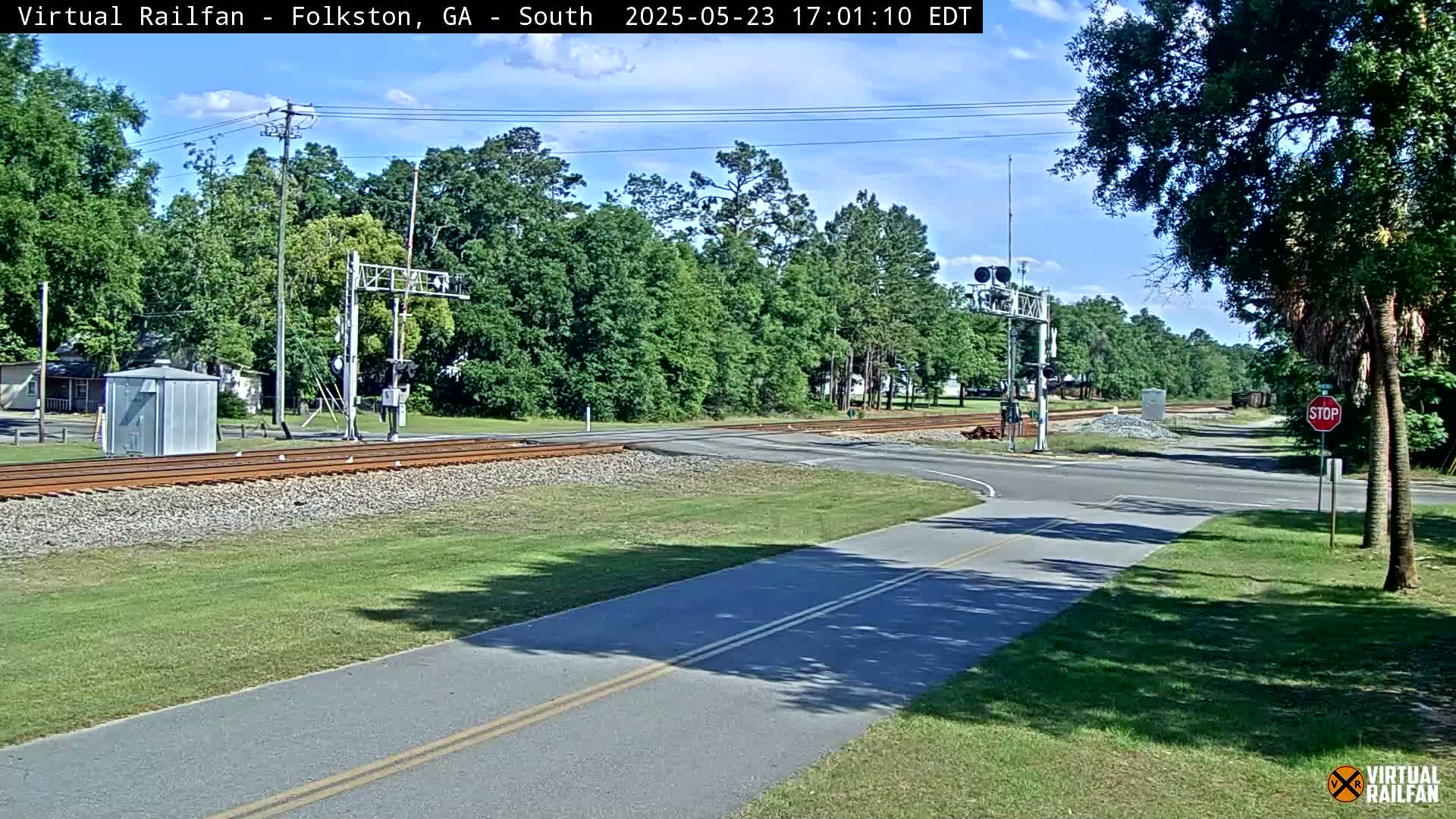 A railroad crossing with a paved road and a bike path intersects under a partly cloudy sky, surrounded by lush green trees.