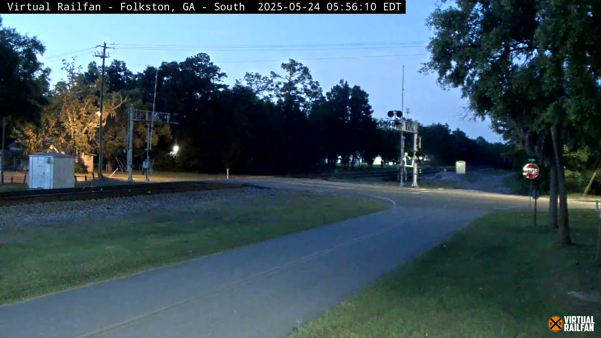 A paved road curves past a railway crossing at dusk, surrounded by trees and under a clear, twilight sky.