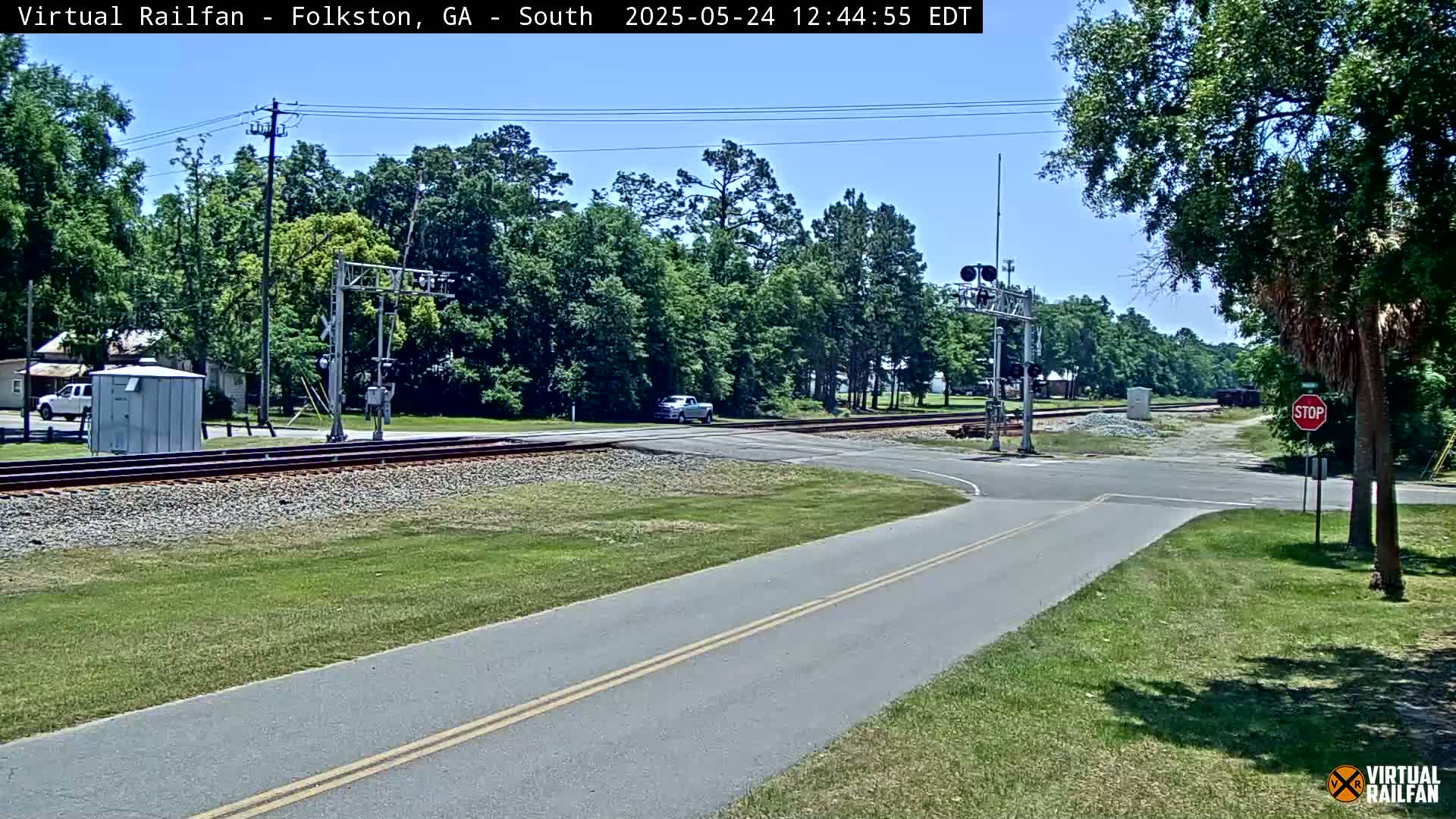 A sunny day reveals a road intersecting with railroad tracks, surrounded by lush green trees and grass, and a vehicle is visible crossing the tracks.