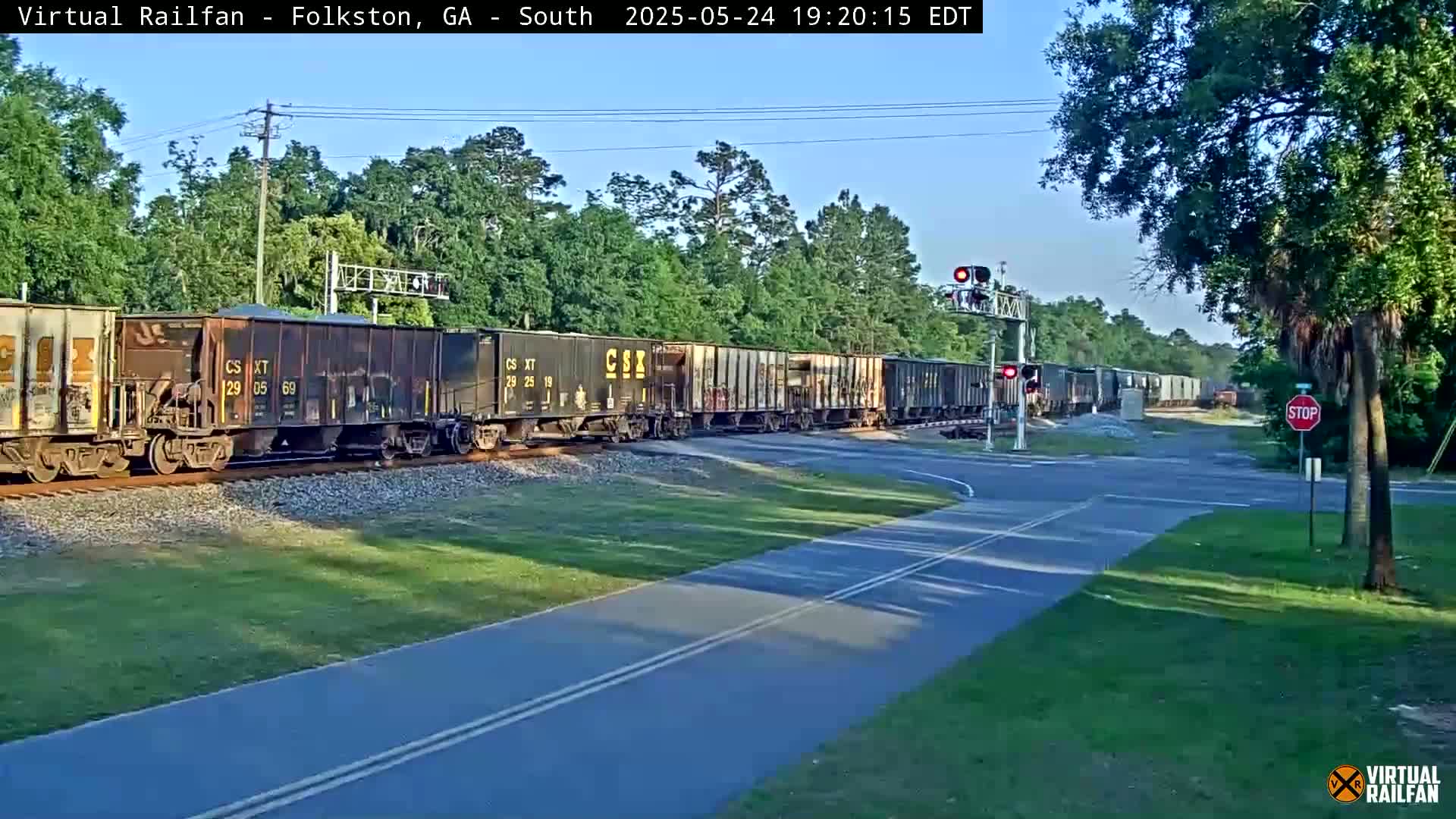 A long freight train passes a road crossing under a clear evening sky, with trees lining the background.