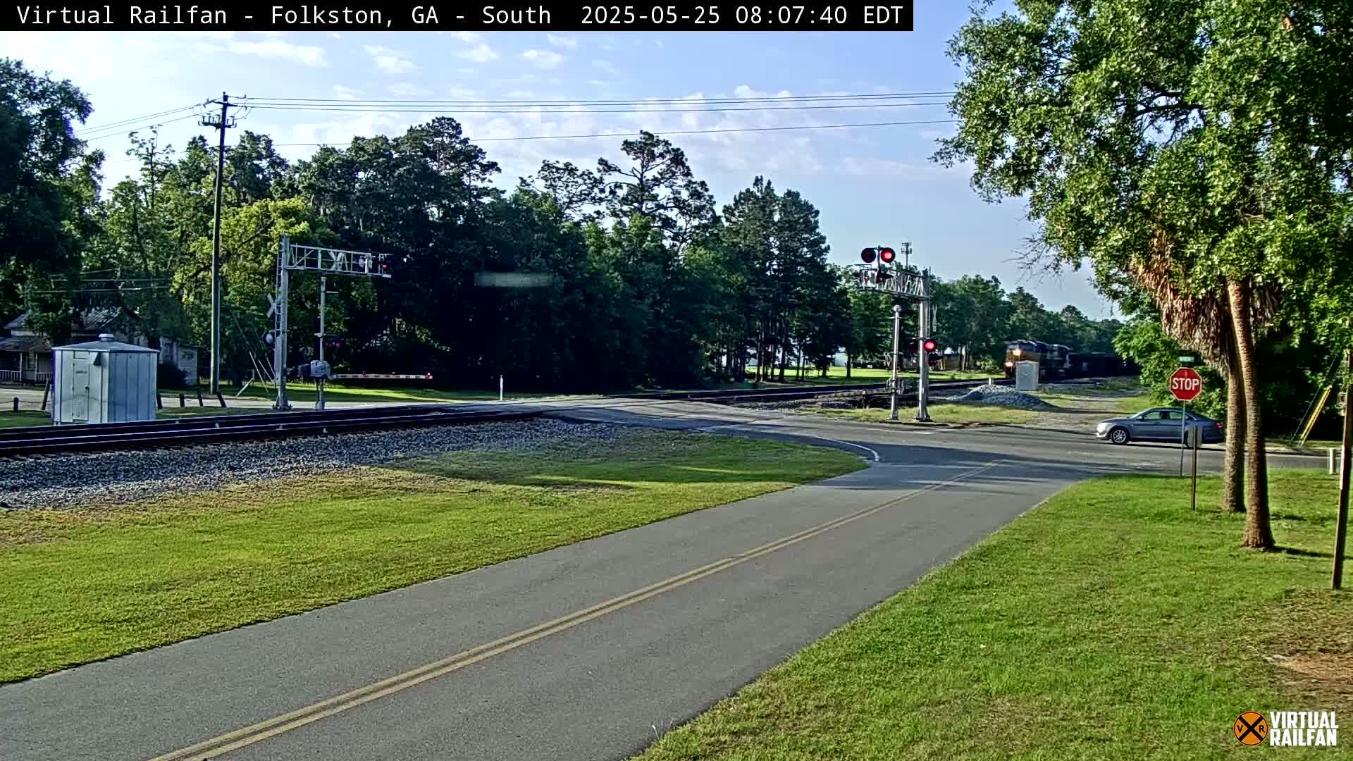 A partly sunny day shows a railroad crossing with a train passing, a car stopped at a stop sign, and a paved road intersecting the tracks.