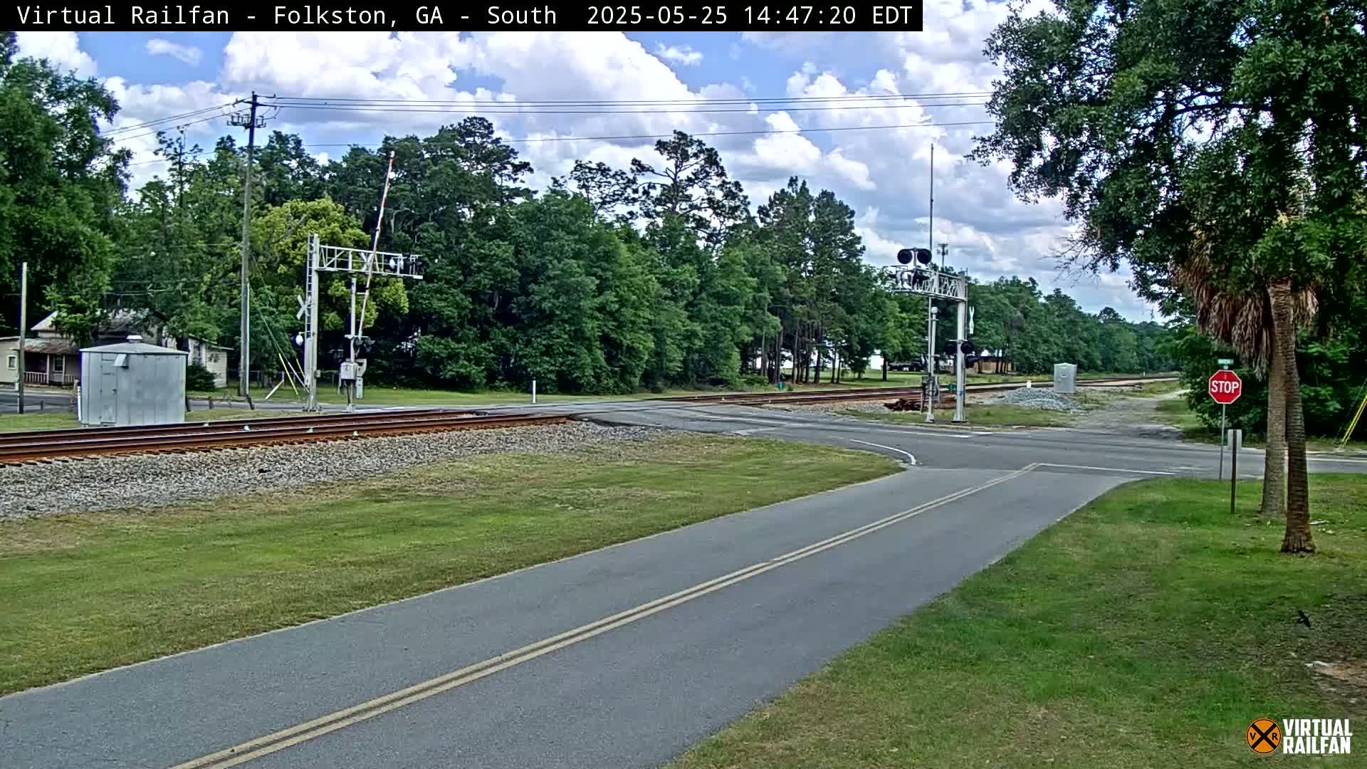 A mostly sunny day shows a two-lane road intersecting a railroad crossing with a stop sign and train signals, surrounded by green grass and trees.