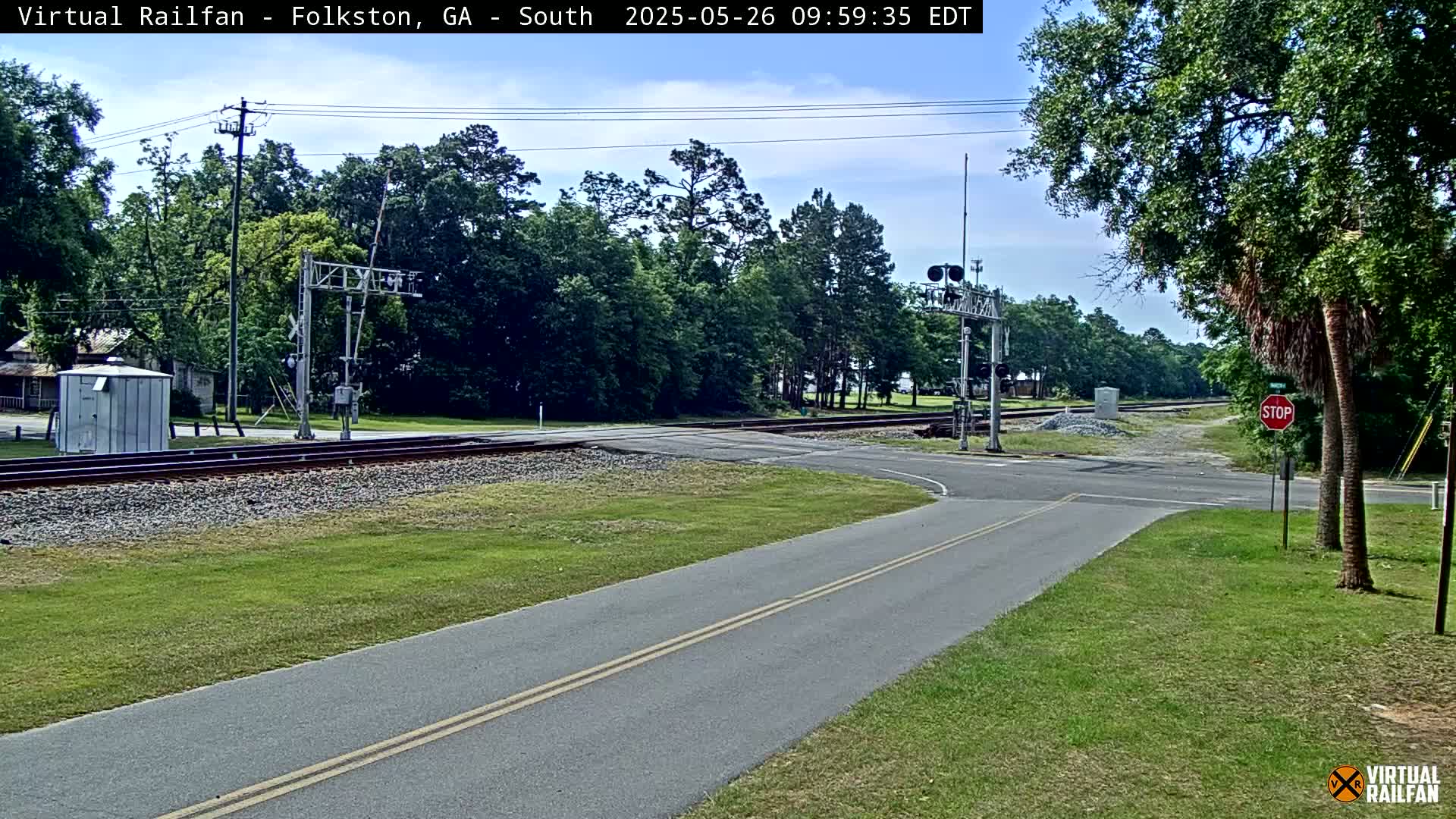 A paved road intersects with a railroad crossing under a partly cloudy sky, surrounded by green vegetation and trees.