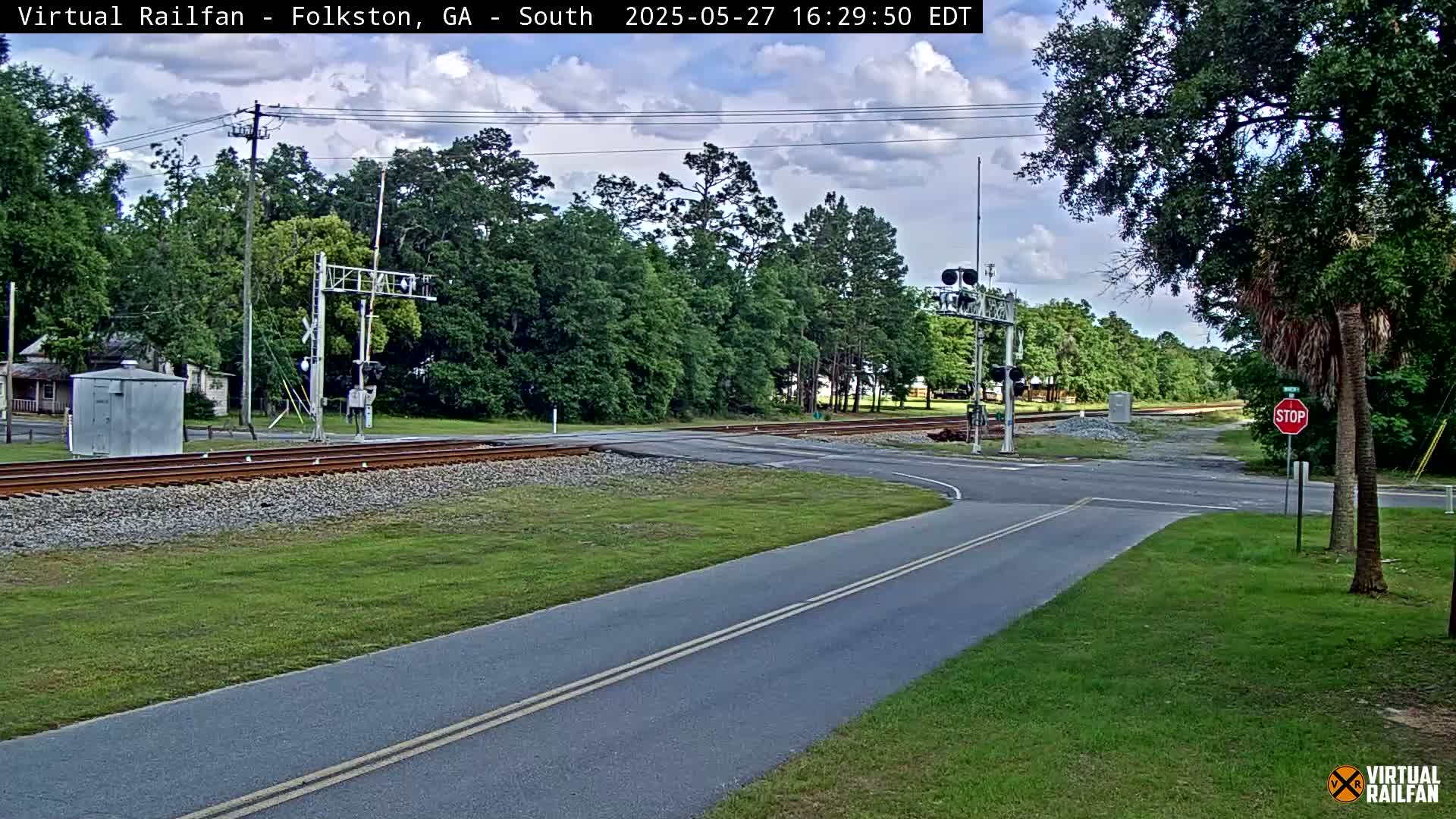 A partly cloudy day shows a railroad crossing intersecting a two-lane road, with a stop sign and lush green vegetation surrounding the scene.