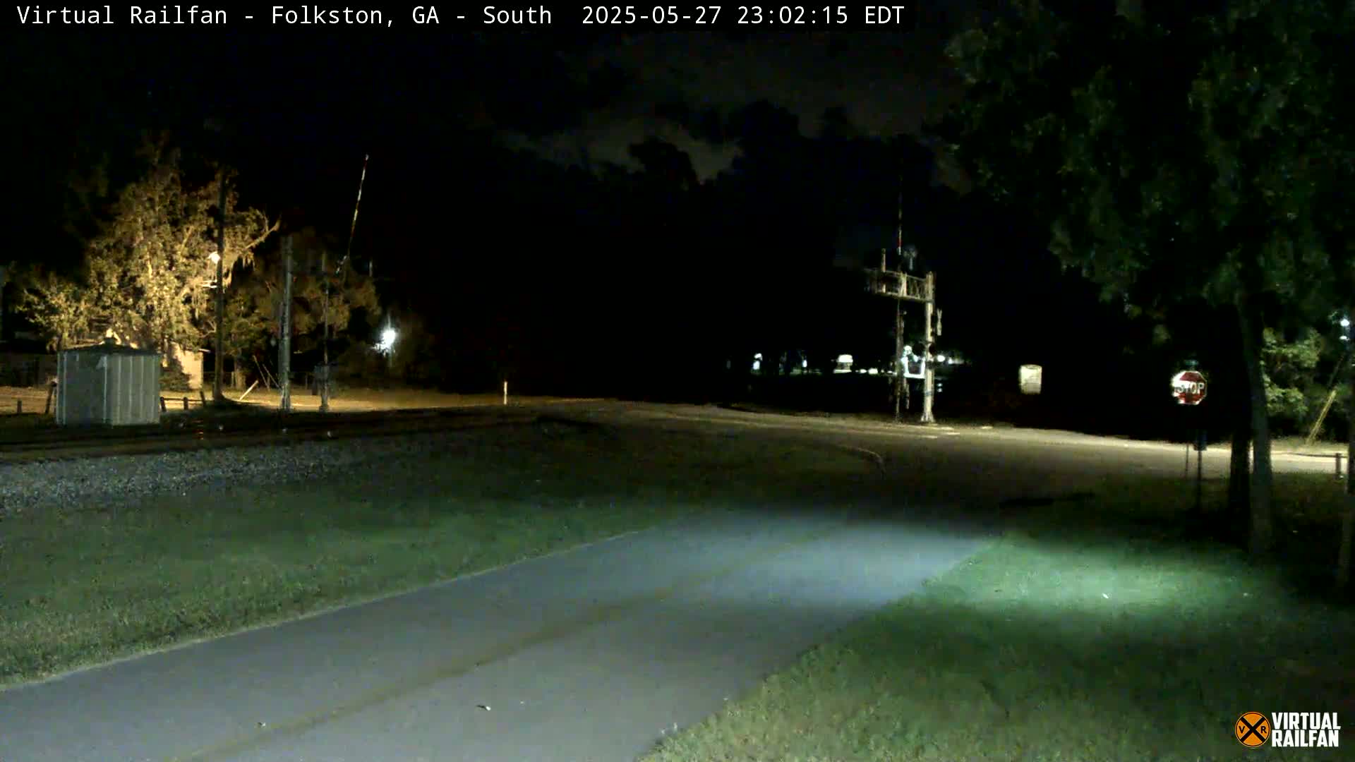 A nighttime view of a paved path intersecting a road near railroad tracks, under a clear sky.