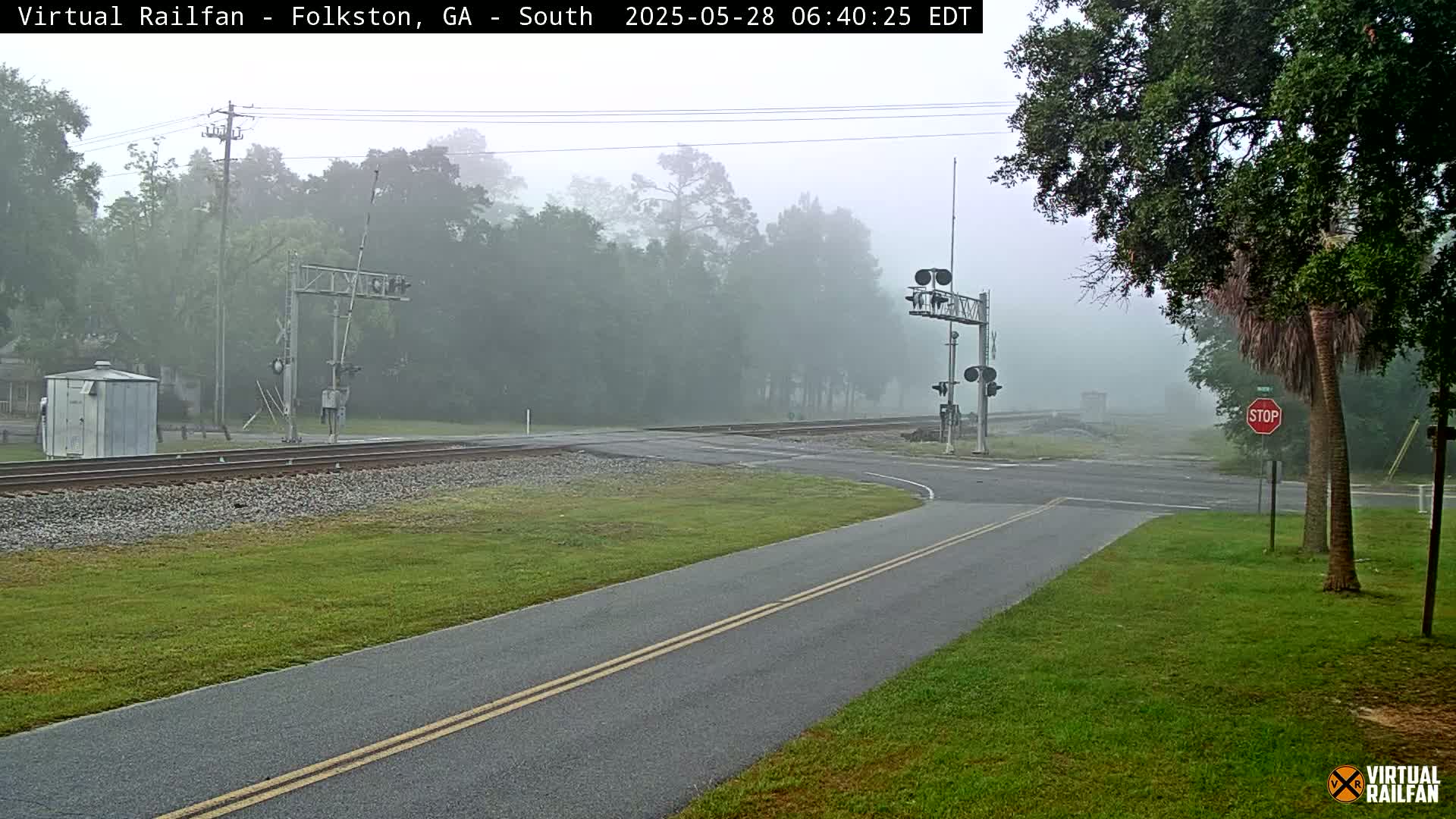 A foggy morning scene shows a railway crossing with a road intersecting the tracks, surrounded by lush green grass and trees.