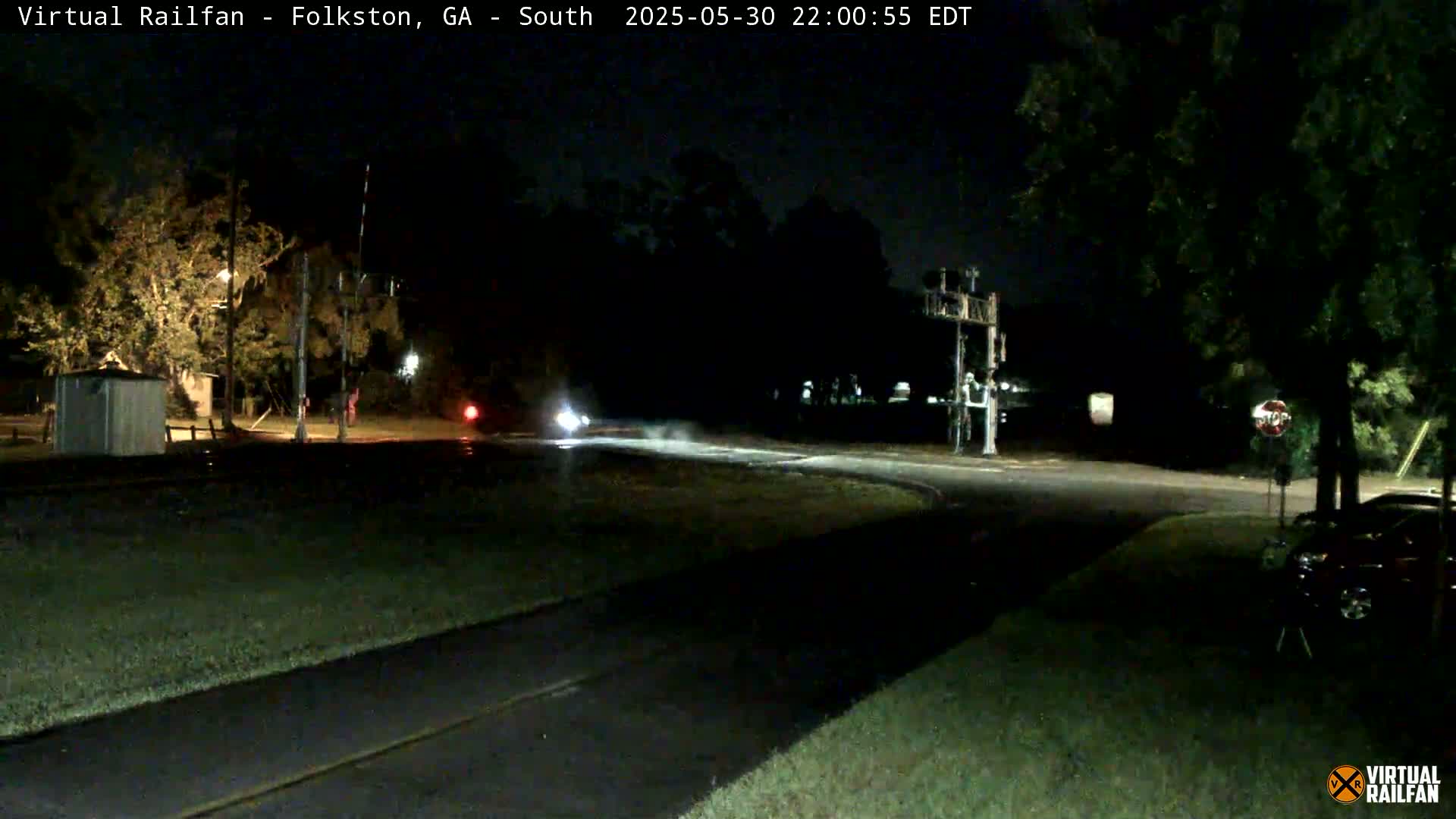 A nighttime view of a rural road intersection with a railroad crossing, showing a few vehicles and some trees under a dark sky.