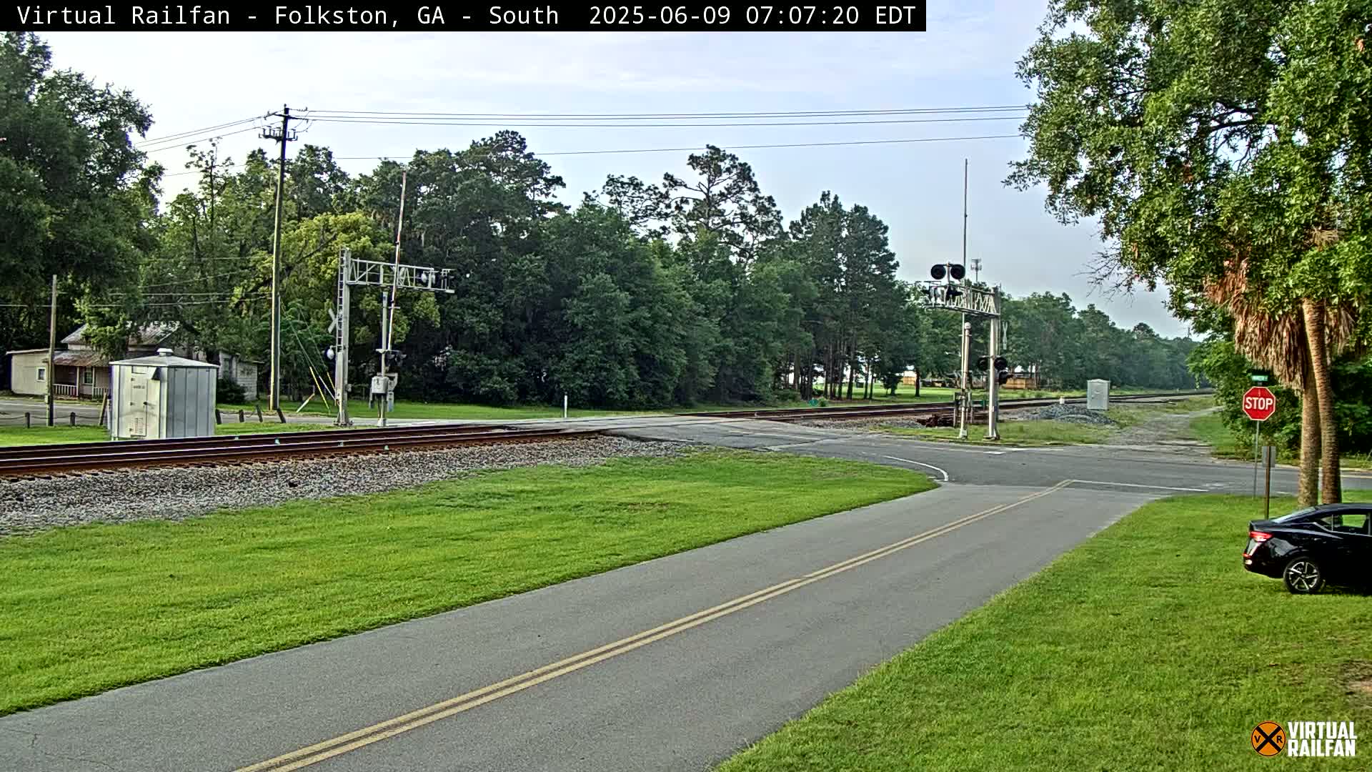A road intersects a railroad crossing under a partly cloudy sky, with lush green grass and trees surrounding the area.