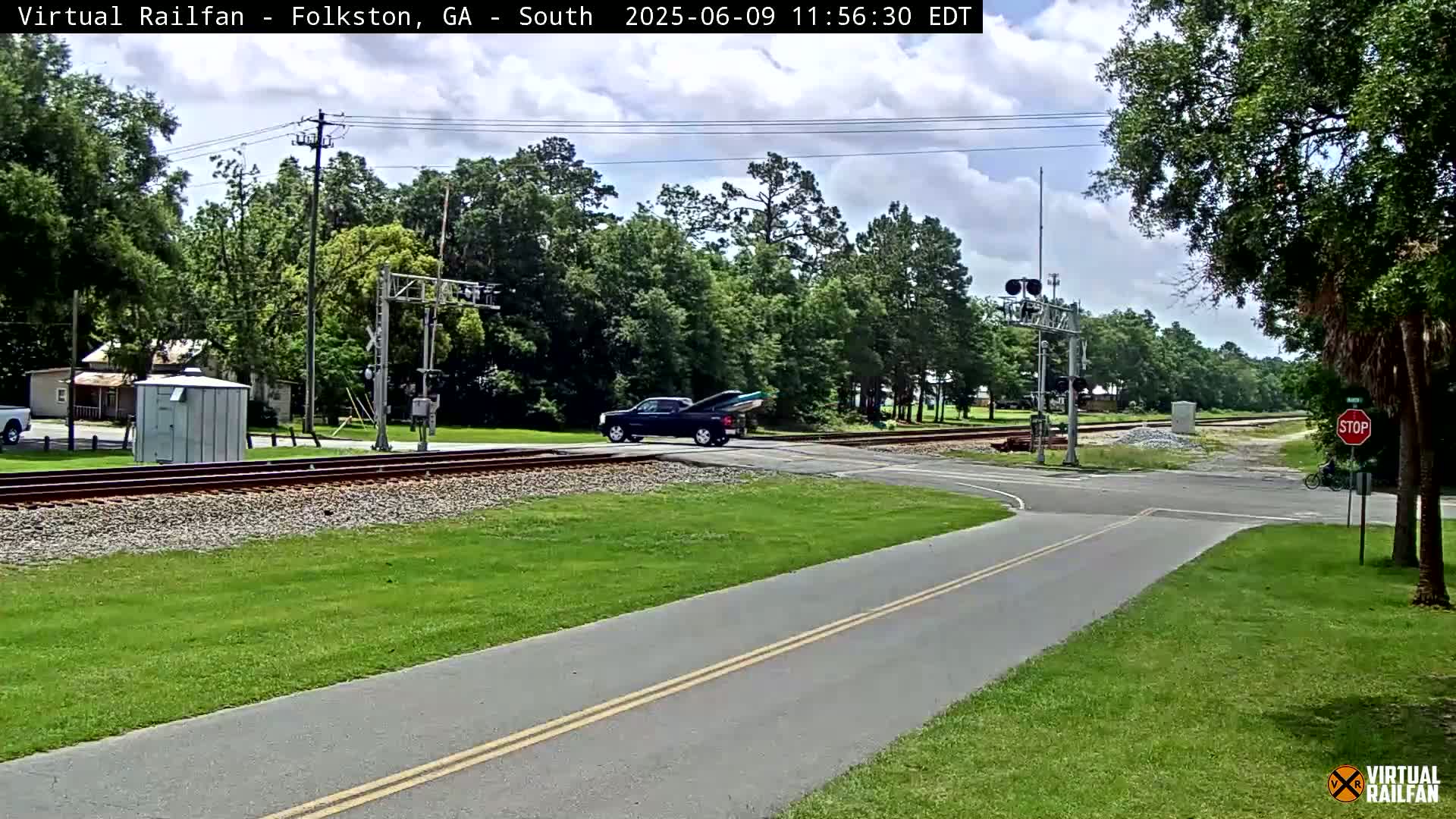 A partly cloudy day shows a dark-colored pickup truck with a boat in its bed crossing a railroad track at a road intersection, surrounded by lush green grass and trees.