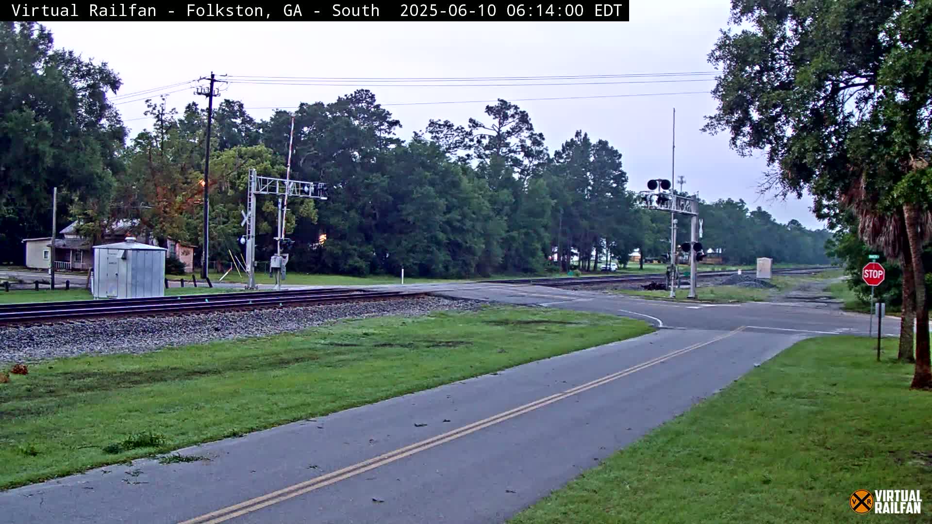 A paved road intersects with railroad tracks at a crossing with train signals and a stop sign, under an overcast sky.