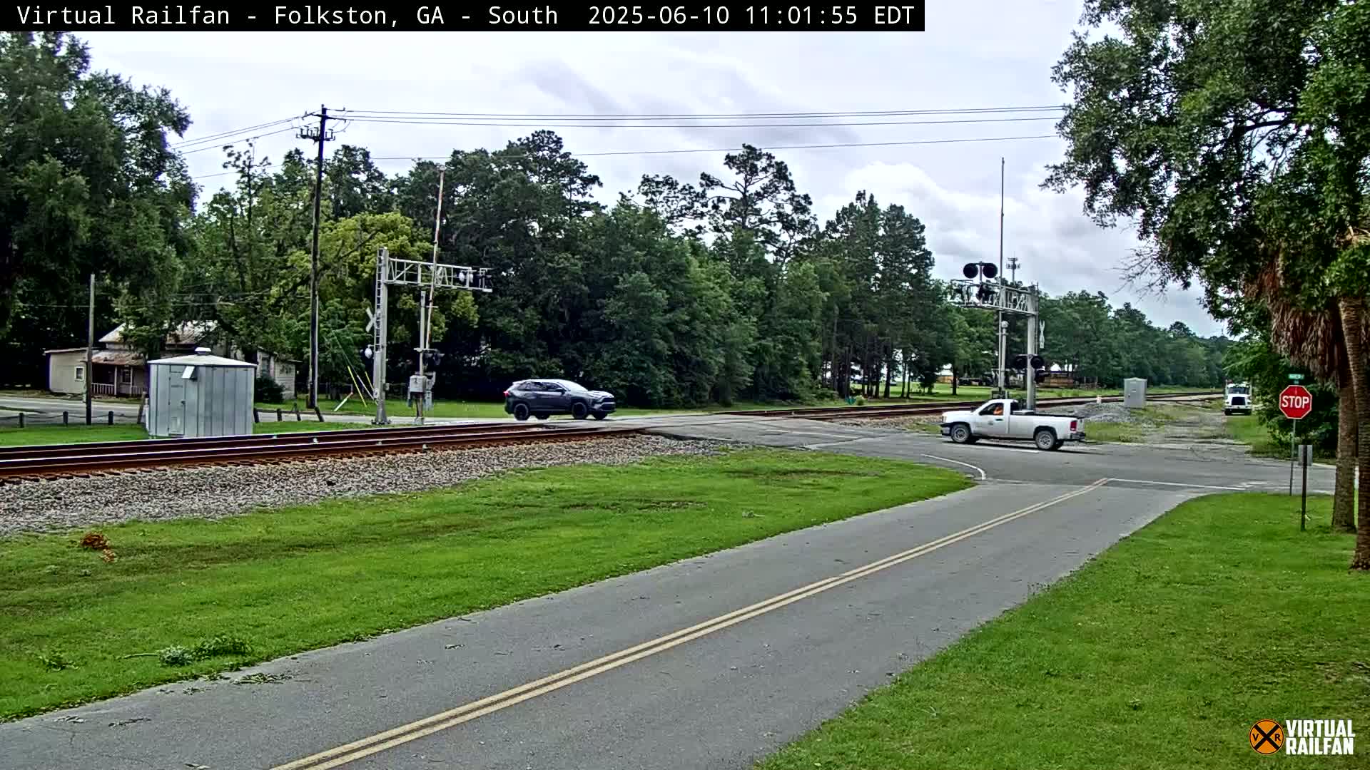 A mostly sunny day shows a road intersecting railroad tracks with a dark SUV and a white pickup truck at the crossing, surrounded by green grass and trees.