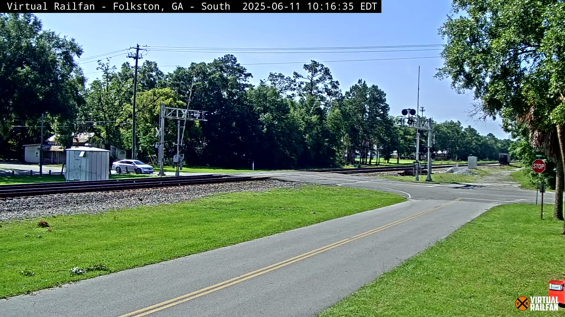 A paved road intersects with railroad tracks at a level crossing under a clear blue sky, surrounded by green grass and lush trees.