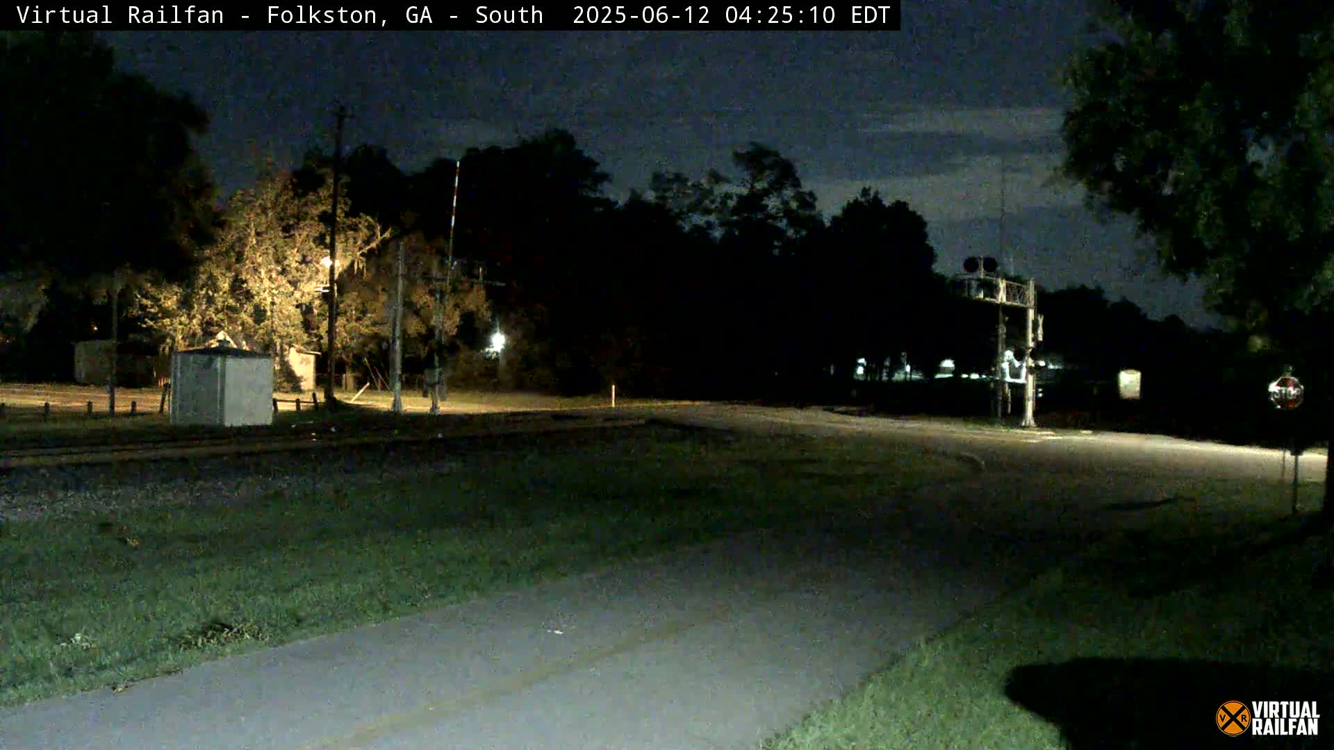 A nighttime view of a railroad crossing with a road intersecting, surrounded by trees under a partly cloudy sky.
